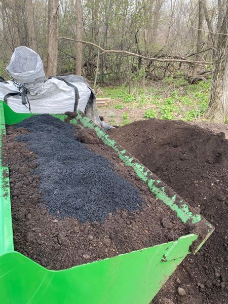 A green tractor bucket holds dark soil and black pellets, near a white bag and trees.