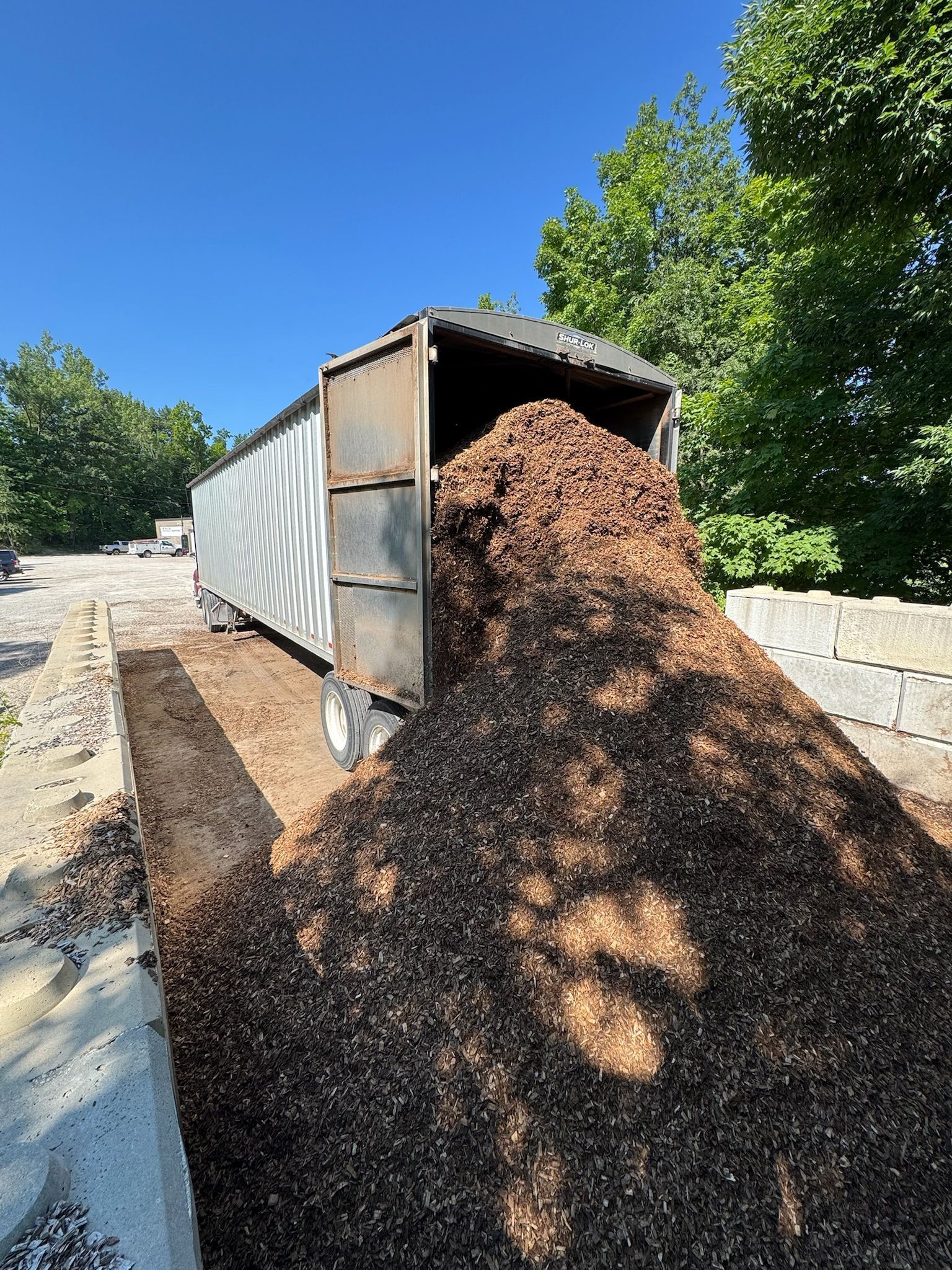 Dump truck unloading a large pile of mulch on a sunny driveway beside a stone wall