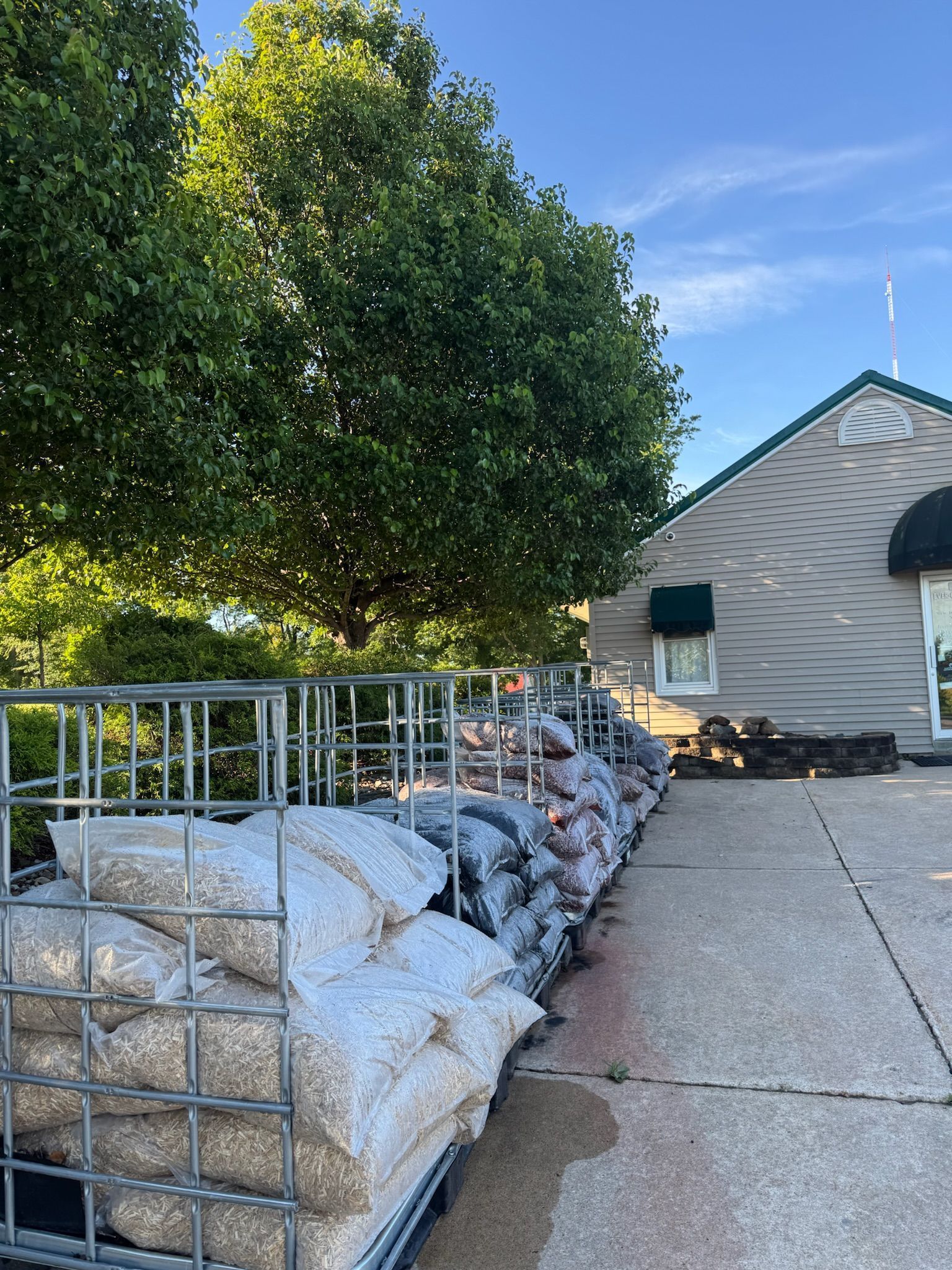 Stacked hay bales beside a driveway and a house, with a large green tree and clear blue sky.