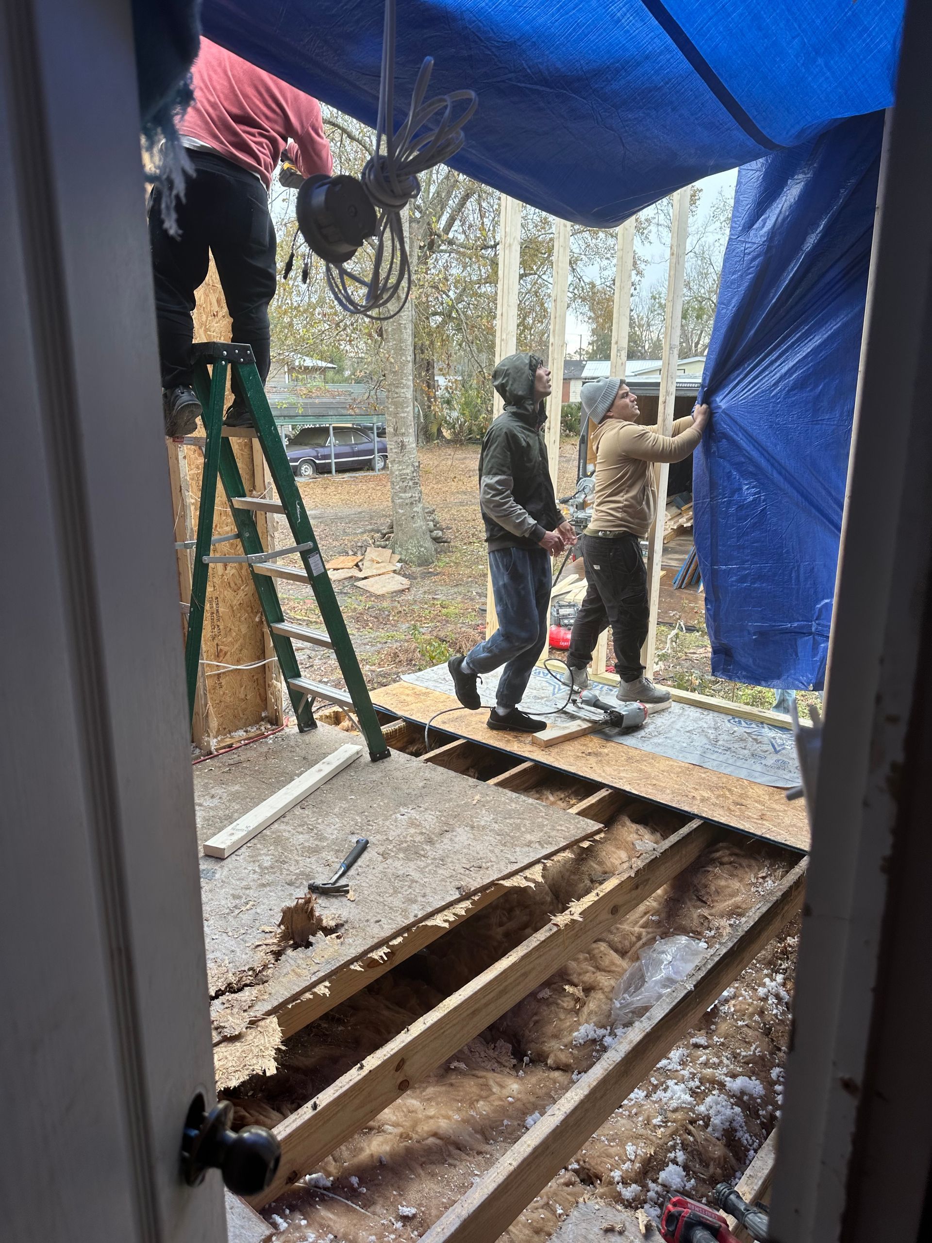 People working on a house frame with a blue tarp. One on ladder, others adjusting tarp; interior view.