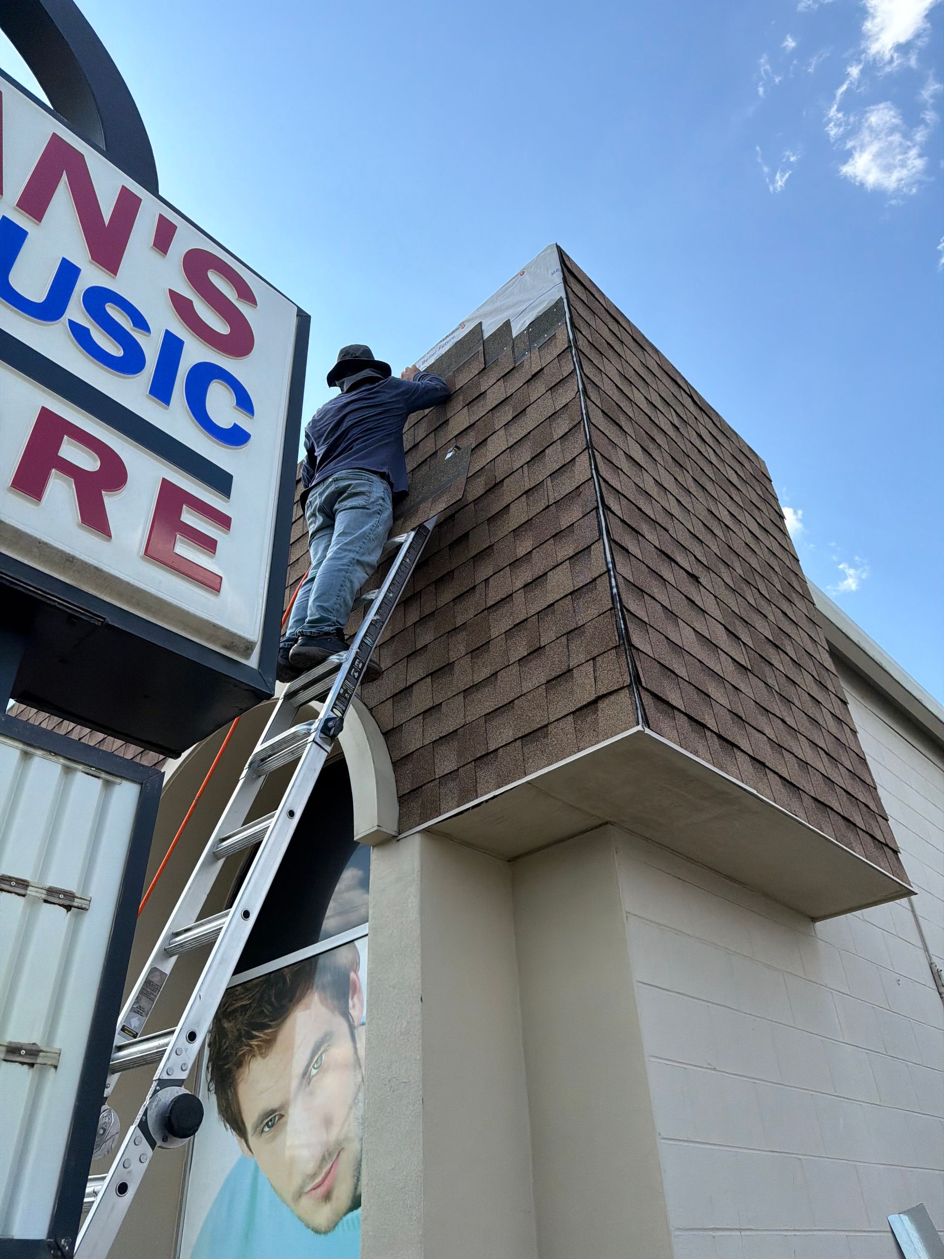 Person on ladder installing shingles on a building's roof, next to a sign for 