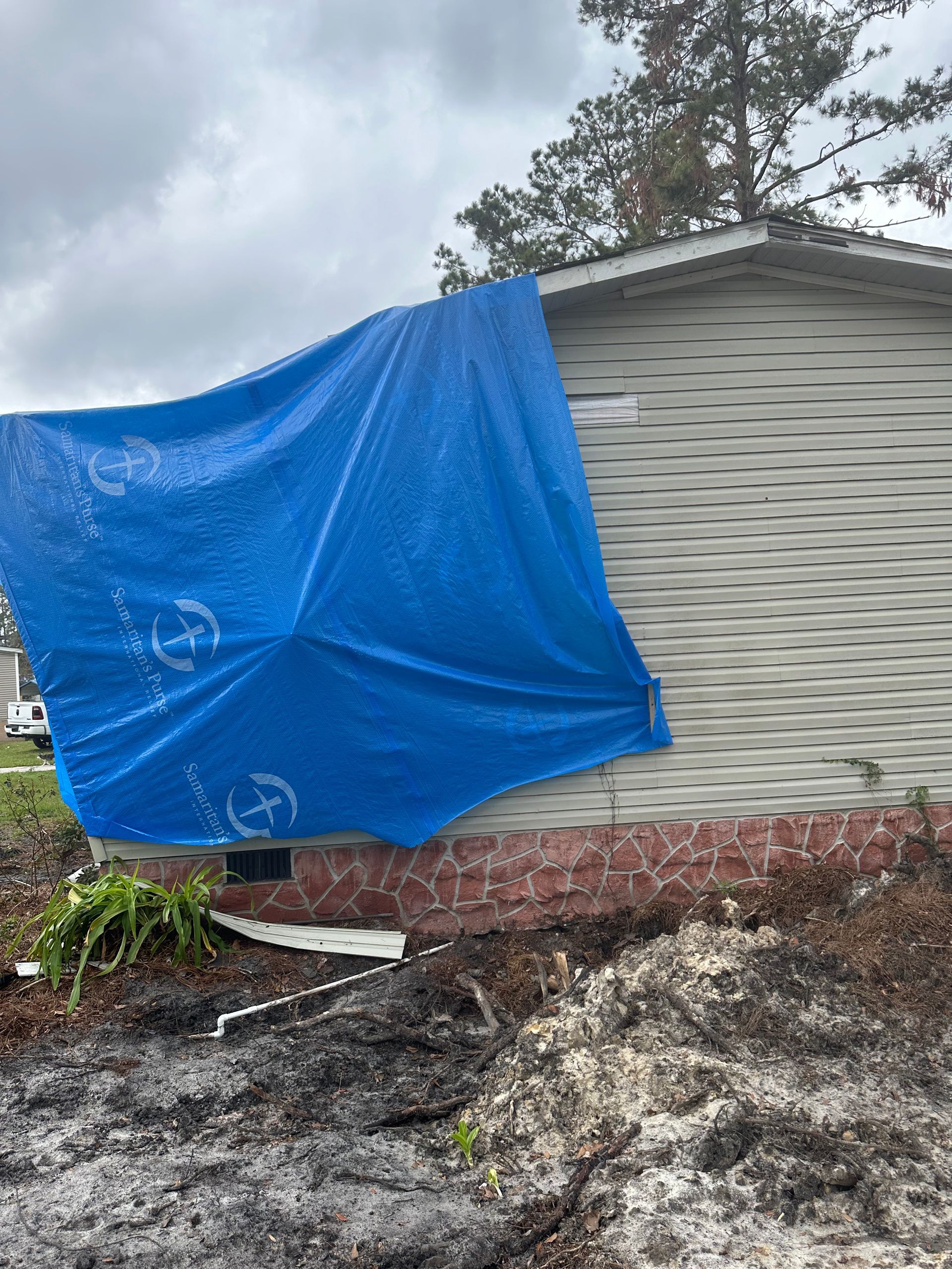 Blue tarp covering damaged side of a house. Green siding, red brick foundation, cloudy sky.