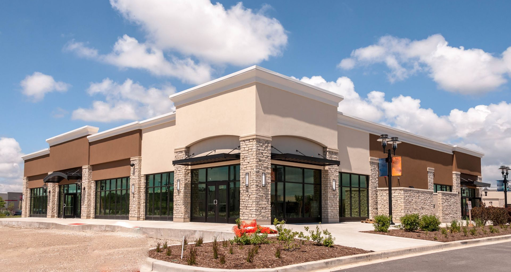 Modern stone-and-brick storefront building with large windows and a landscaped entrance under a blue sky
