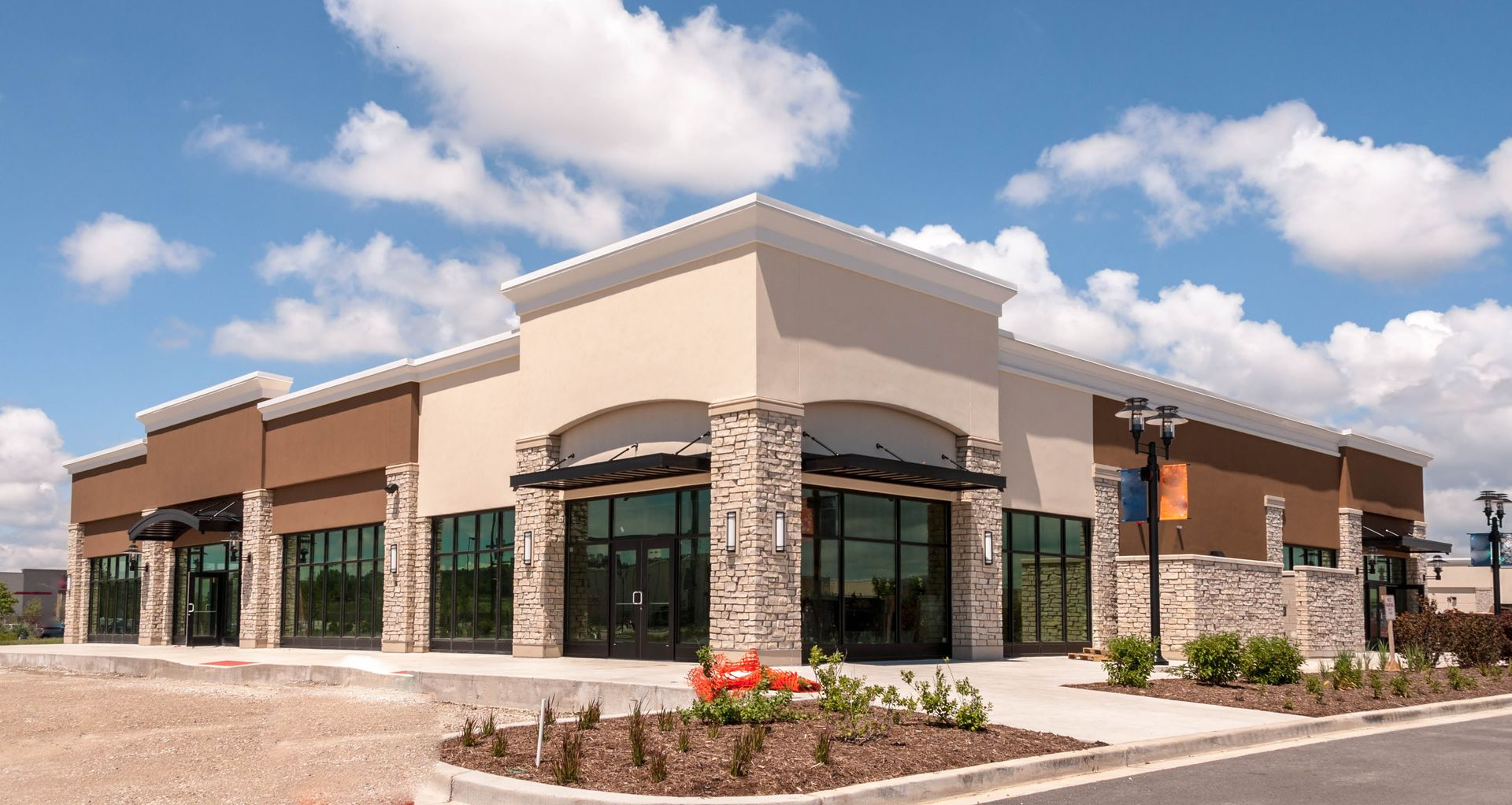 Modern stone-and-brick storefront building with large windows and a landscaped entrance under a blue sky