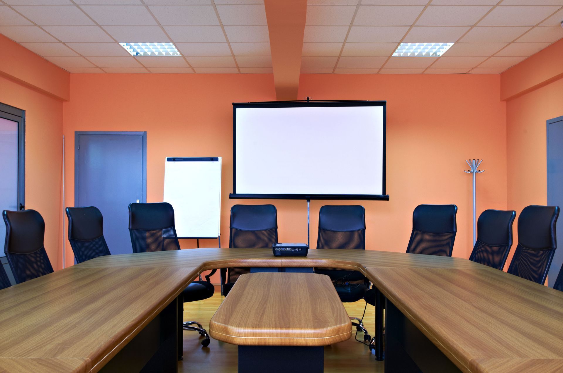 Conference room with a U-shaped table, black chairs, and a projection screen on orange walls.