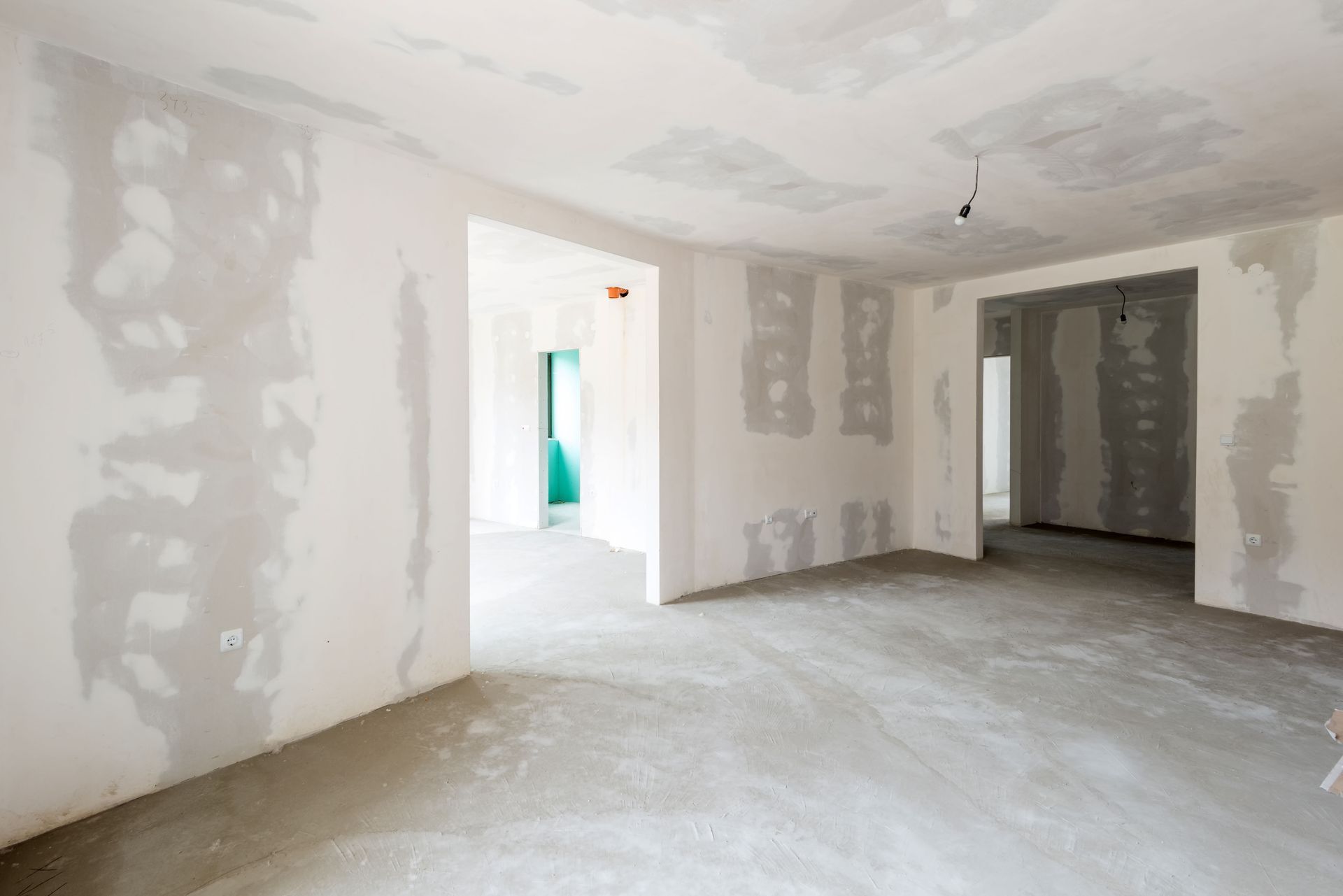 Empty unfinished room with drywall patches, bare floor, and doorways leading to another room.