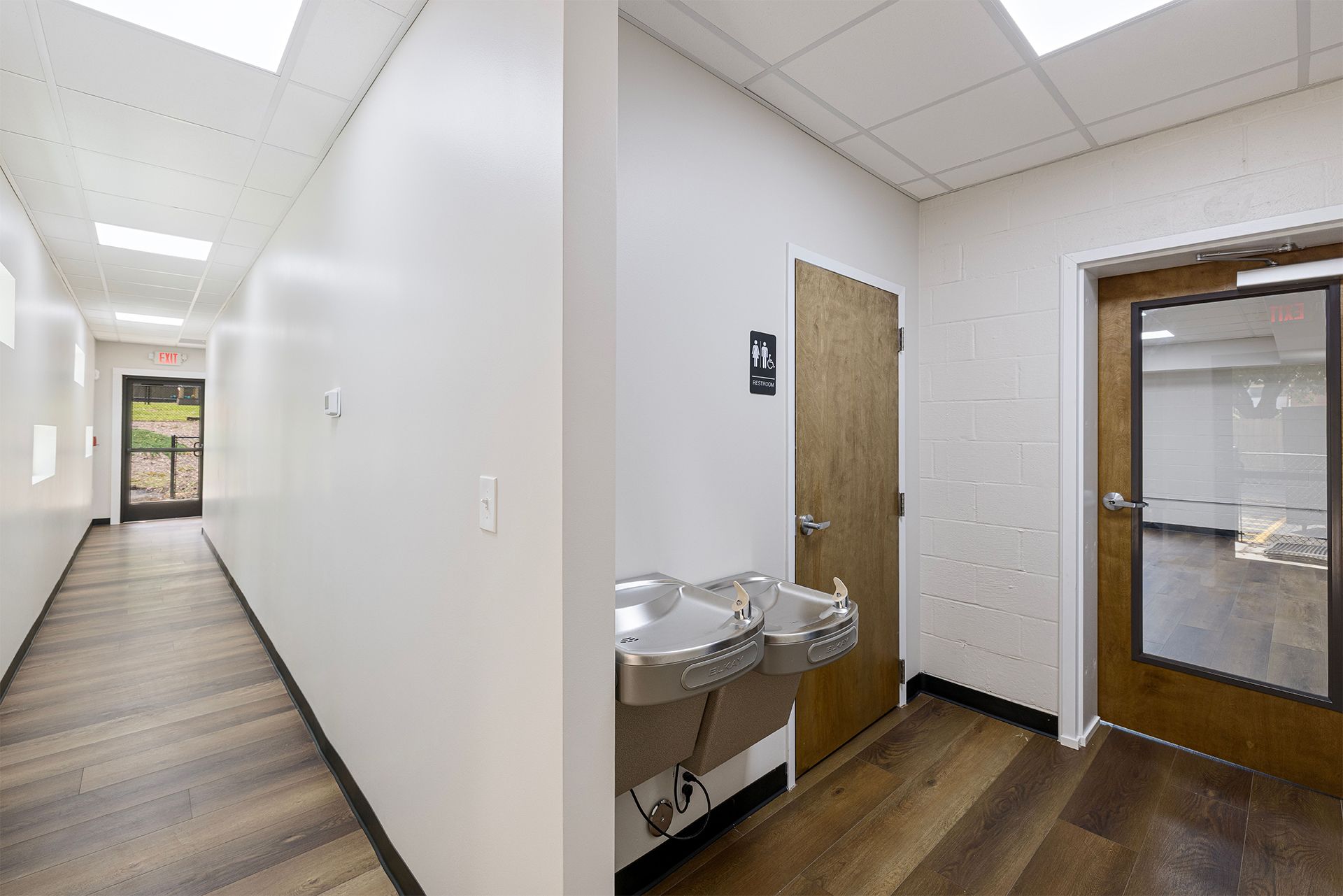 Bright hallway with white walls, wood floor, water fountain, and a door near an adjoining room.