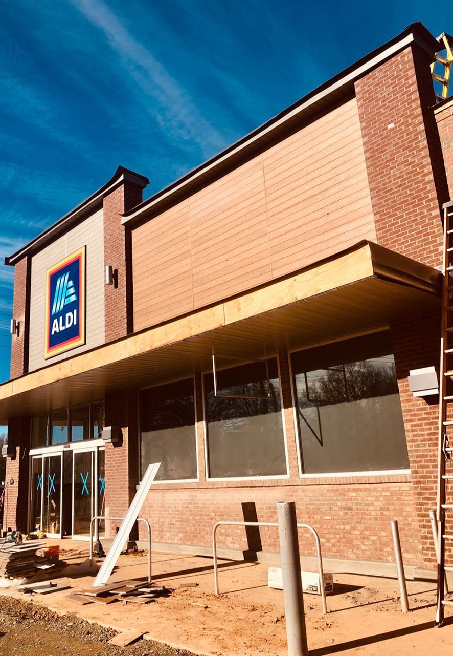 Storefront under construction with tan brick facade, blue sign, and boarded windows under a clear sky