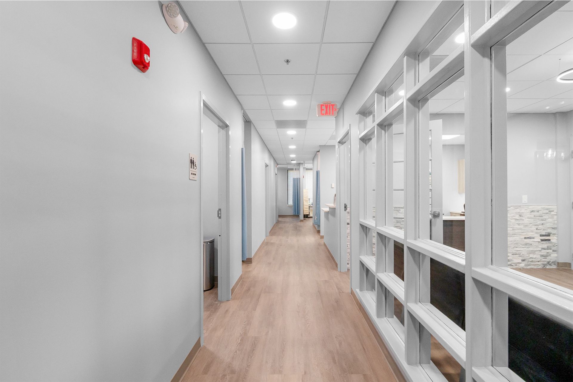 Bright office hallway with wood floors, glass walls, and white doors leading to a distant red exit sign