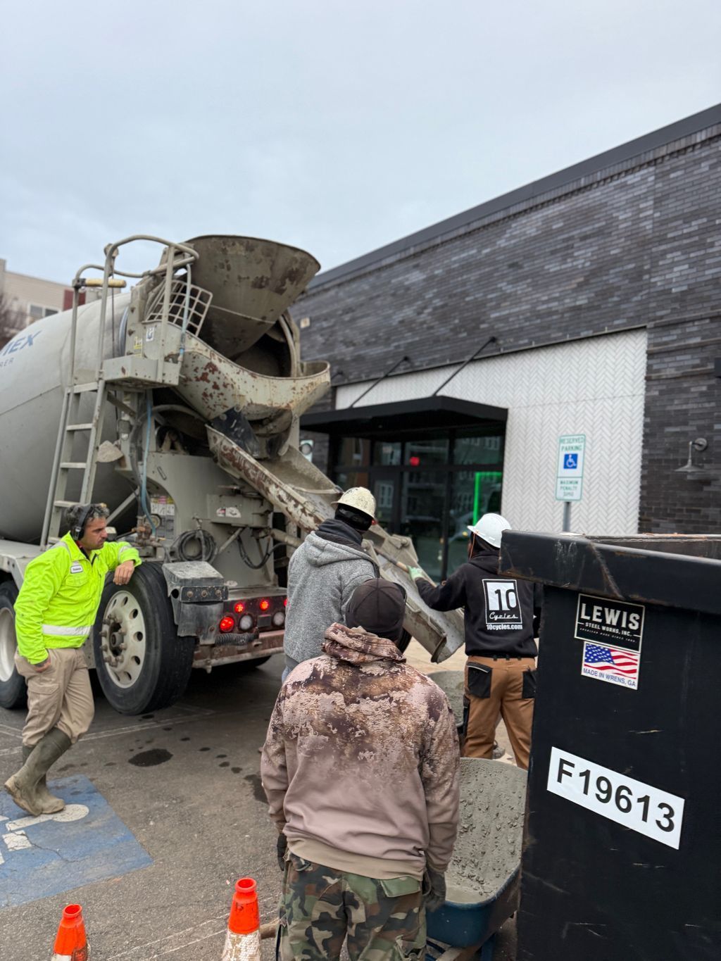Workers guide a cement mixer at a construction site beside a warehouse, with orange cones nearby.