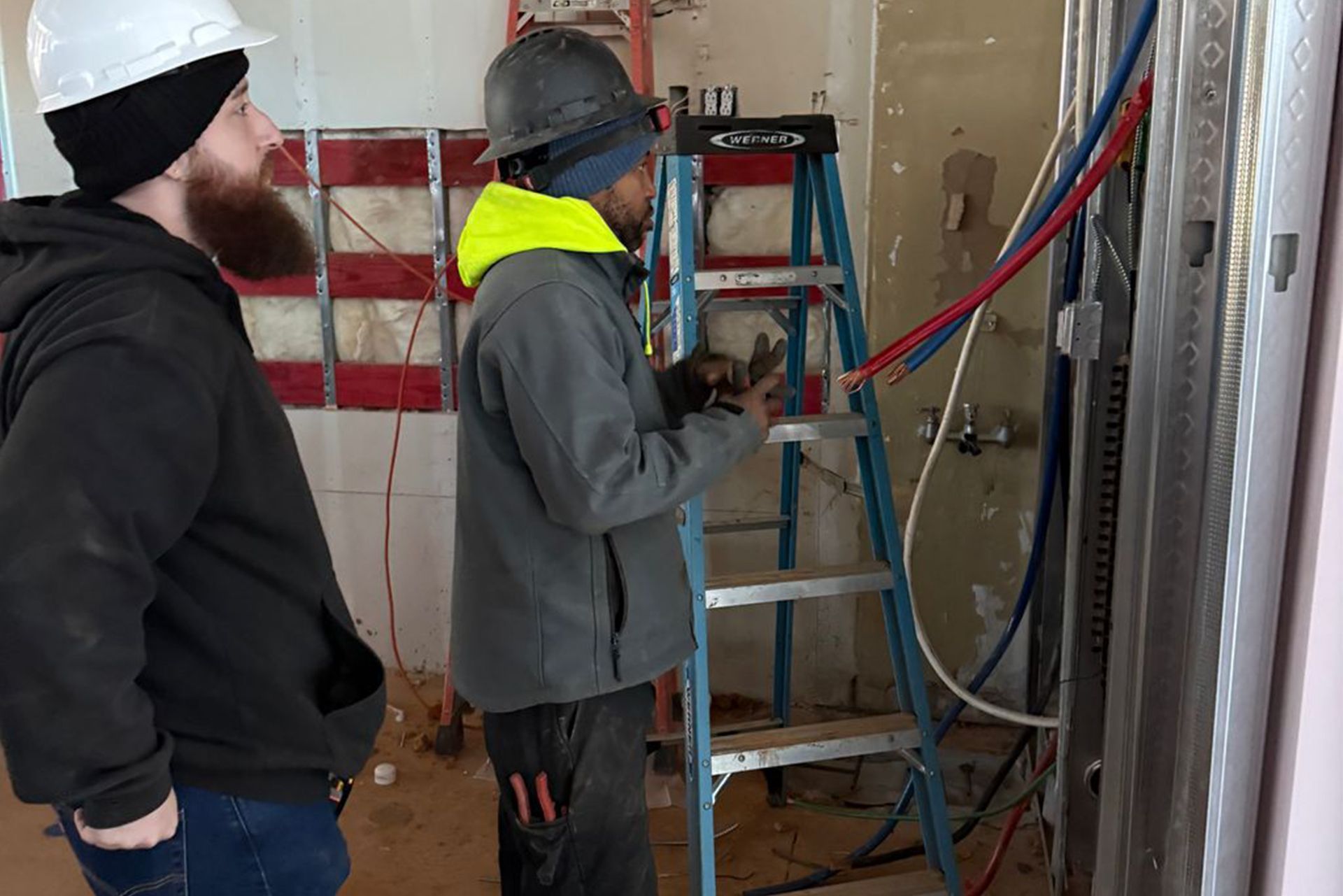Two workers in hard hats install wiring in an unfinished room, with a ladder and exposed studs.