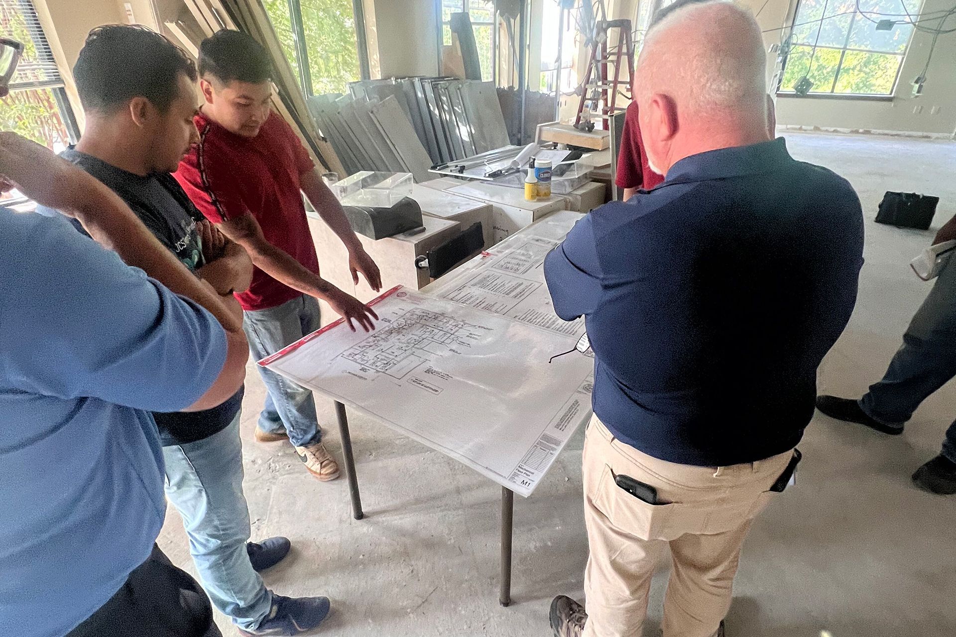 Four people study large floor plans on a table in a bright room with construction materials nearby