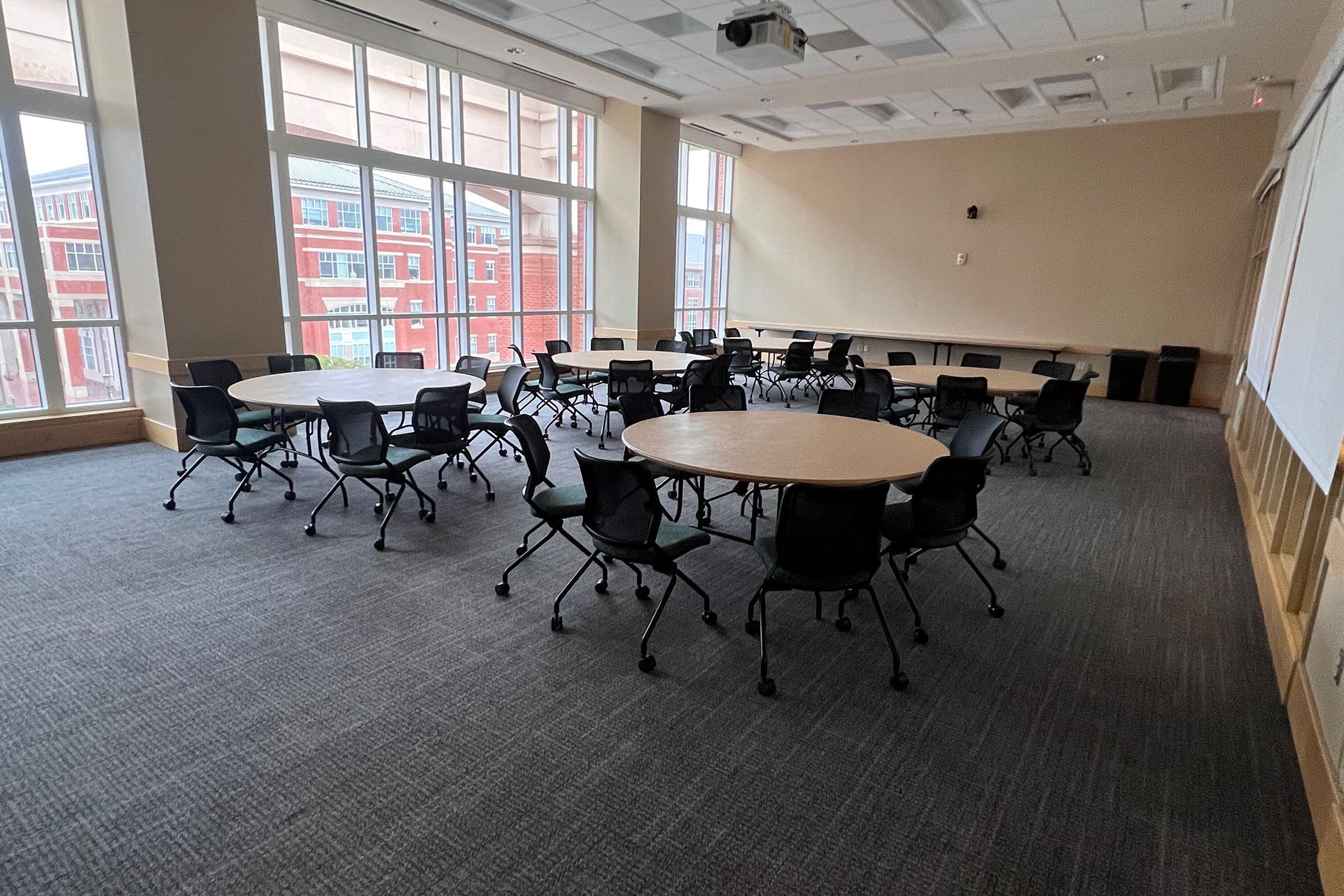 Empty conference room with round tables and black chairs, large windows, and carpeted floor.