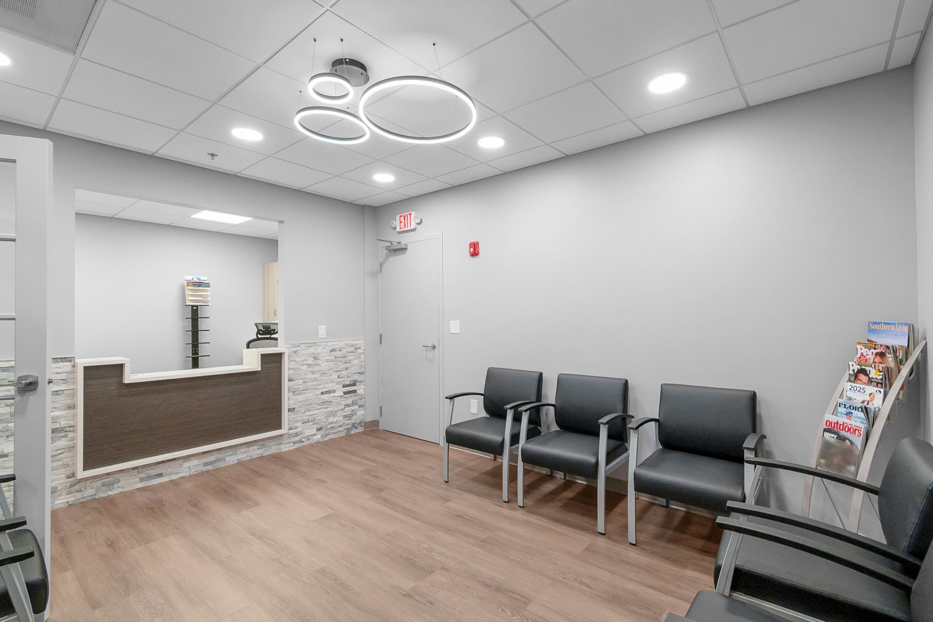 Empty waiting room with gray chairs, wood floor, reception desk, and ceiling ring lights.