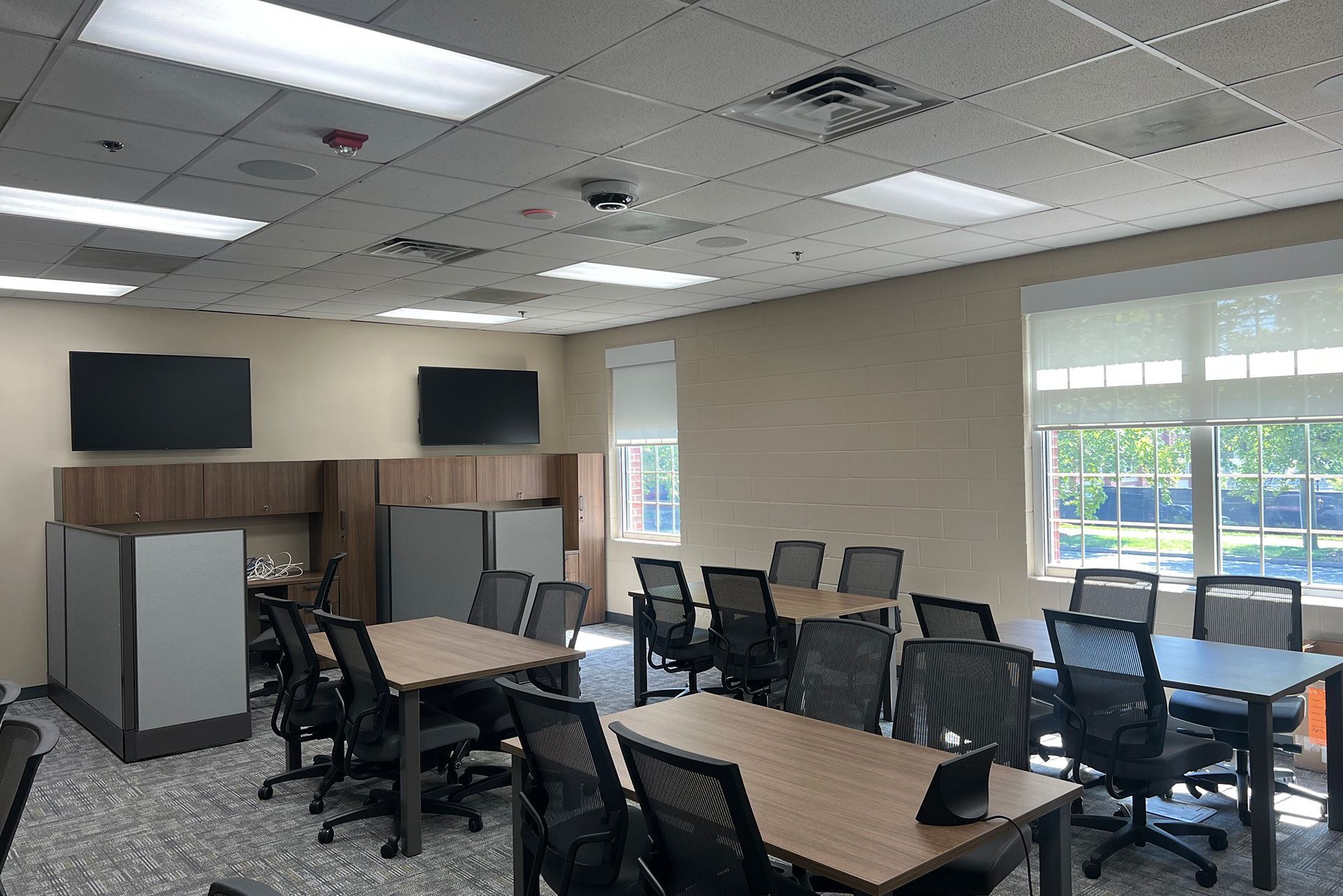 Empty classroom with tables, chairs, wall-mounted screens, and large windows letting in daylight