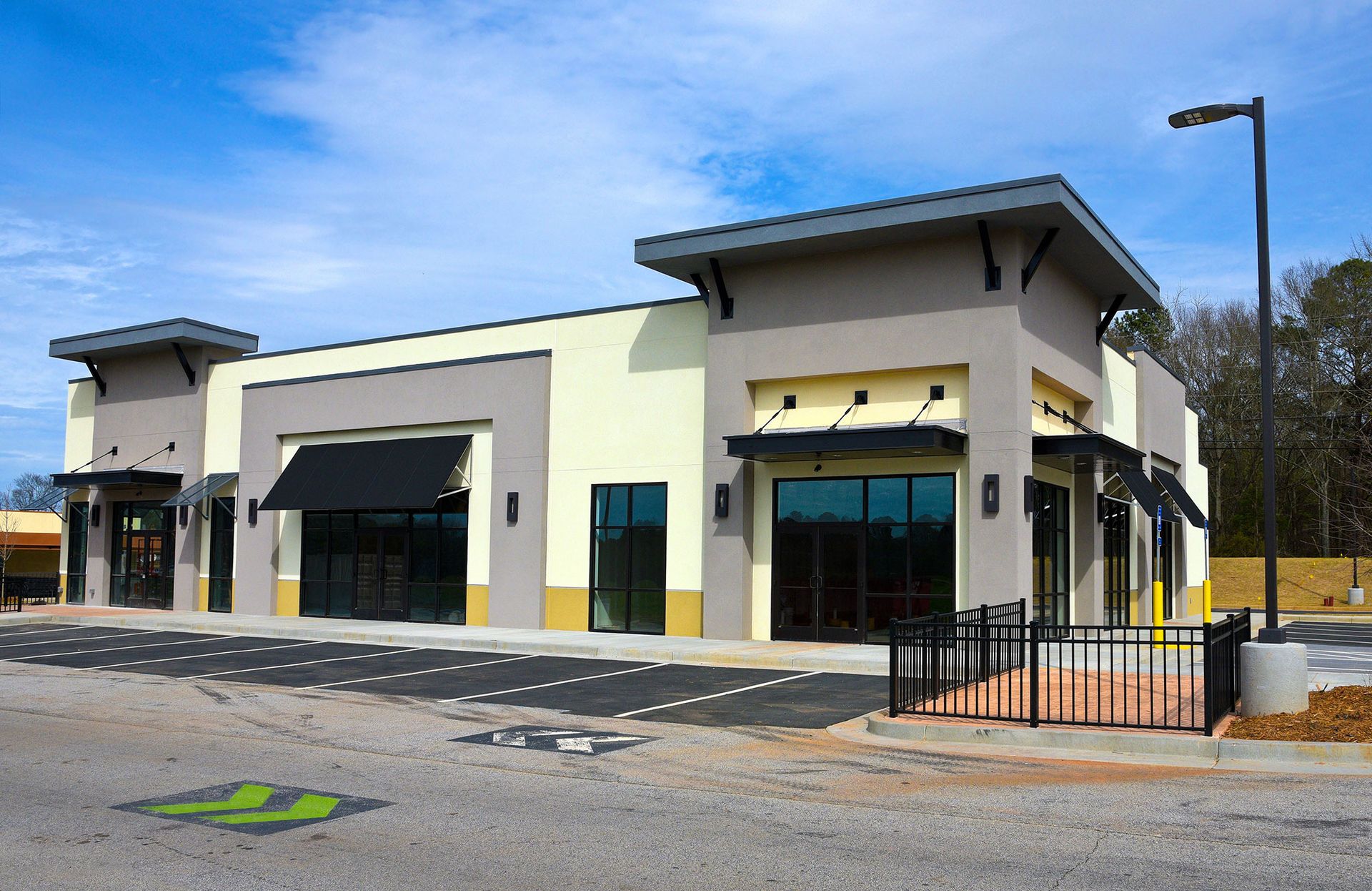 Modern commercial building with large windows, cream and gray exterior, and a small fenced entrance under a blue sky