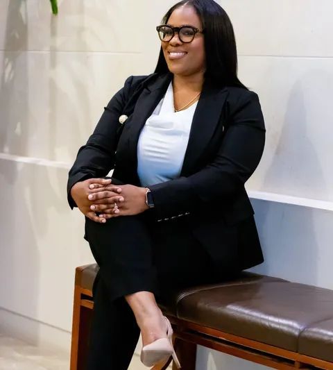 Smiling woman in a black blazer sitting on a bench with hands clasped in a bright indoor lobby