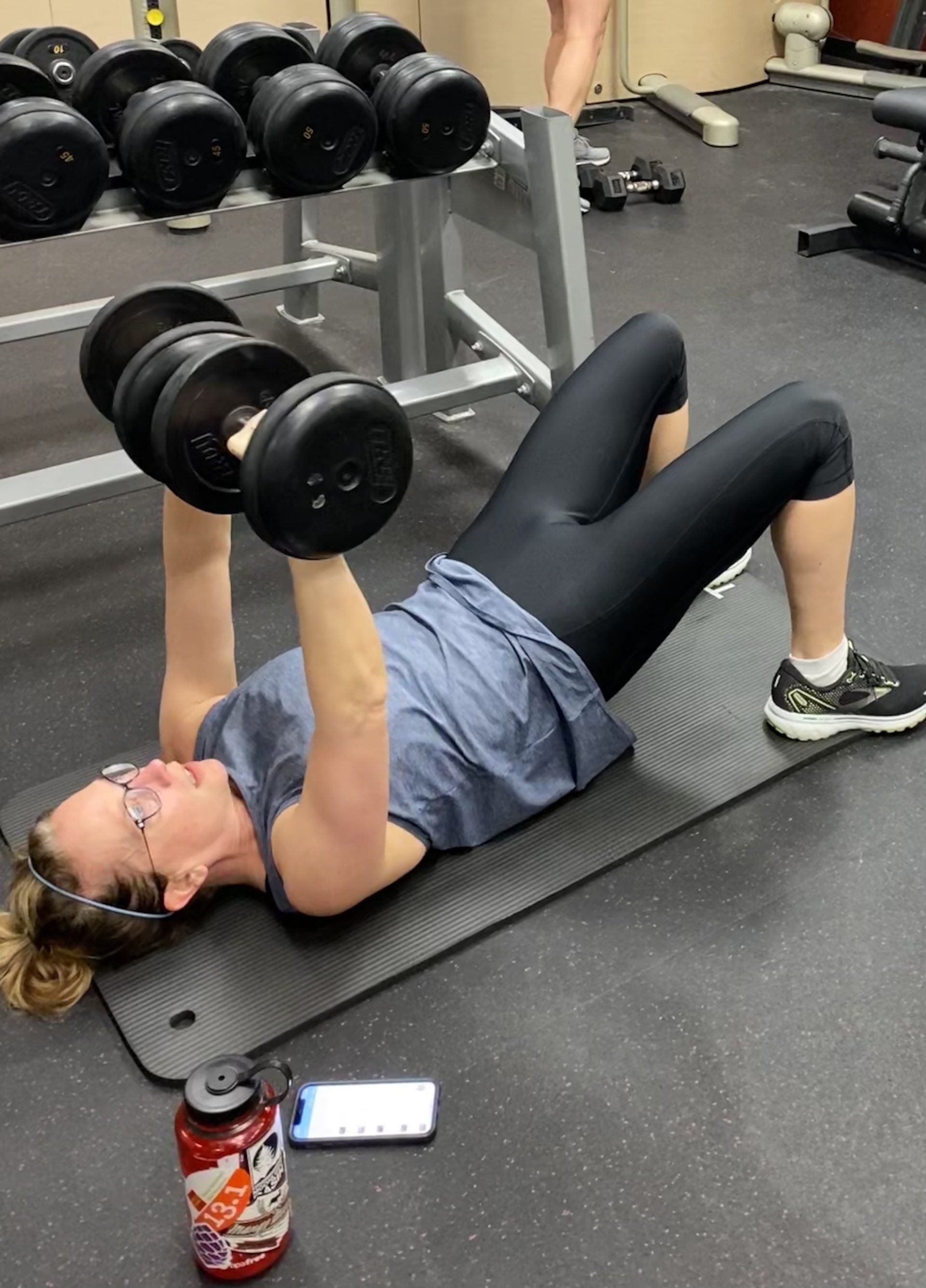 A woman is laying on a mat lifting dumbbells in a gym