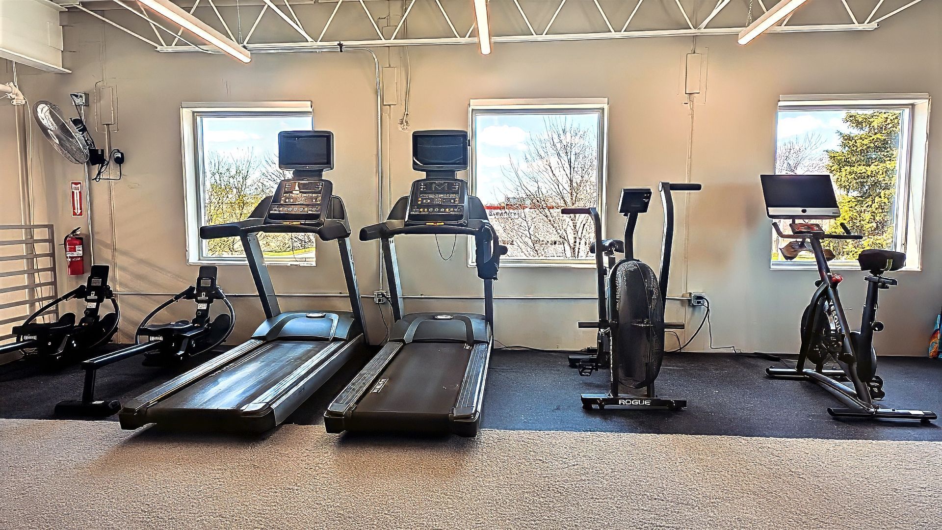 Gym with treadmills, bikes, and weight machines in a bright room with windows.