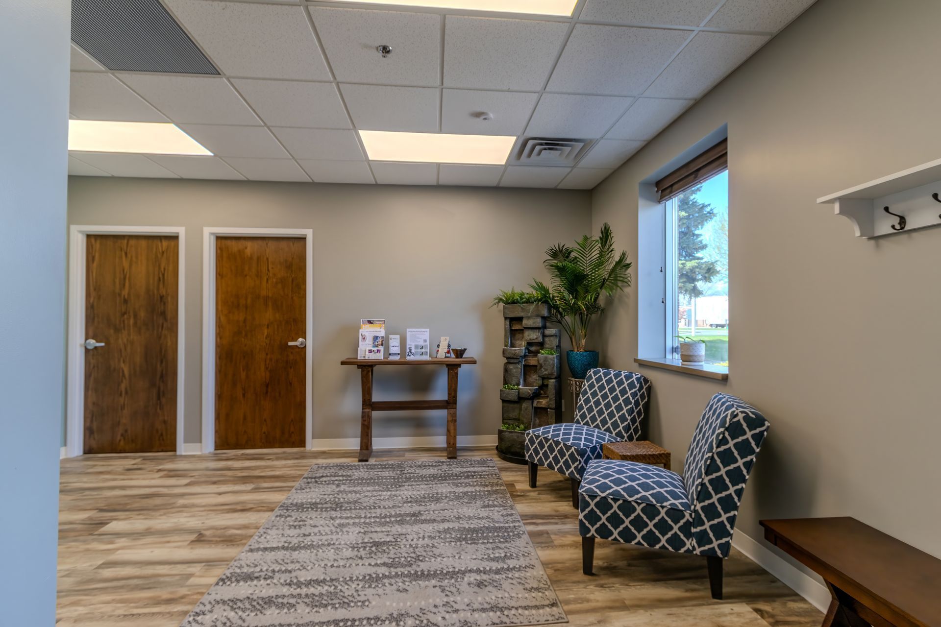 Empty waiting room with two patterned chairs, a small table, wood doors, and a window with a plant.