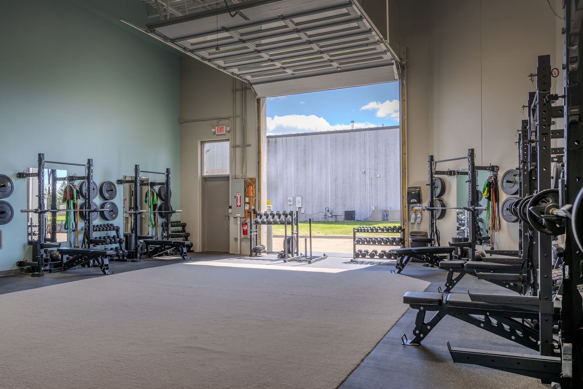 Empty gym interior with weight racks and benches by an open garage door.