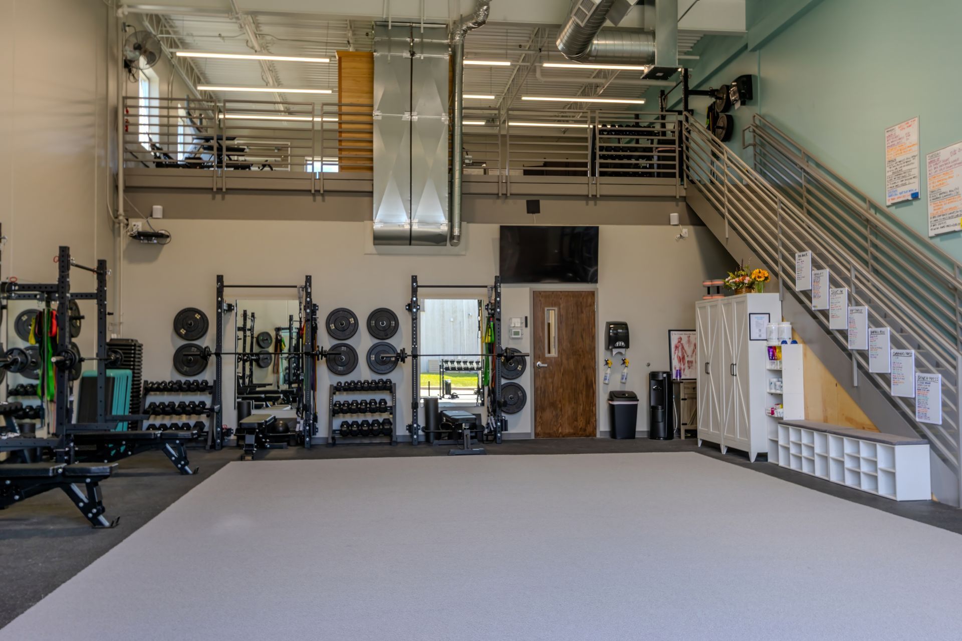 Spacious gym with weight racks, benches, and a staircase leading to an upper mezzanine area.