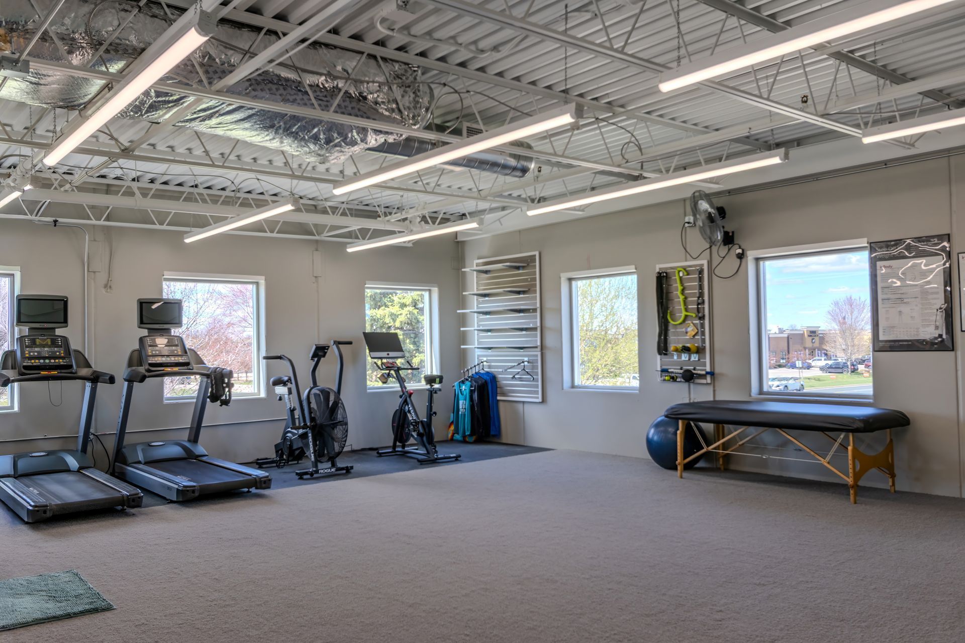 Empty gym room with treadmills, exercise bikes, and large windows letting in daylight.