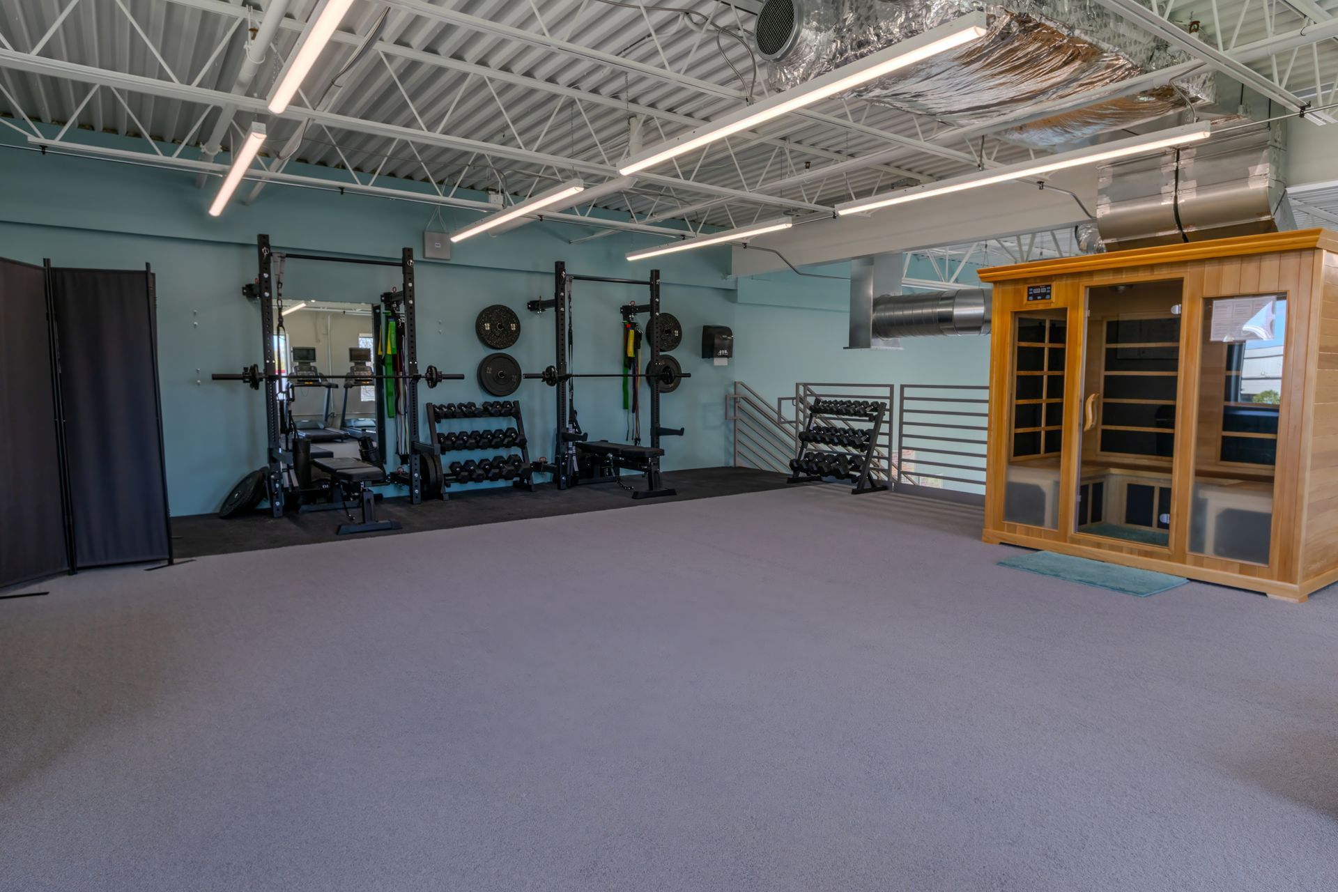 Empty gym studio with carpeted floor, weight racks, mirrors, and a wooden sauna room.