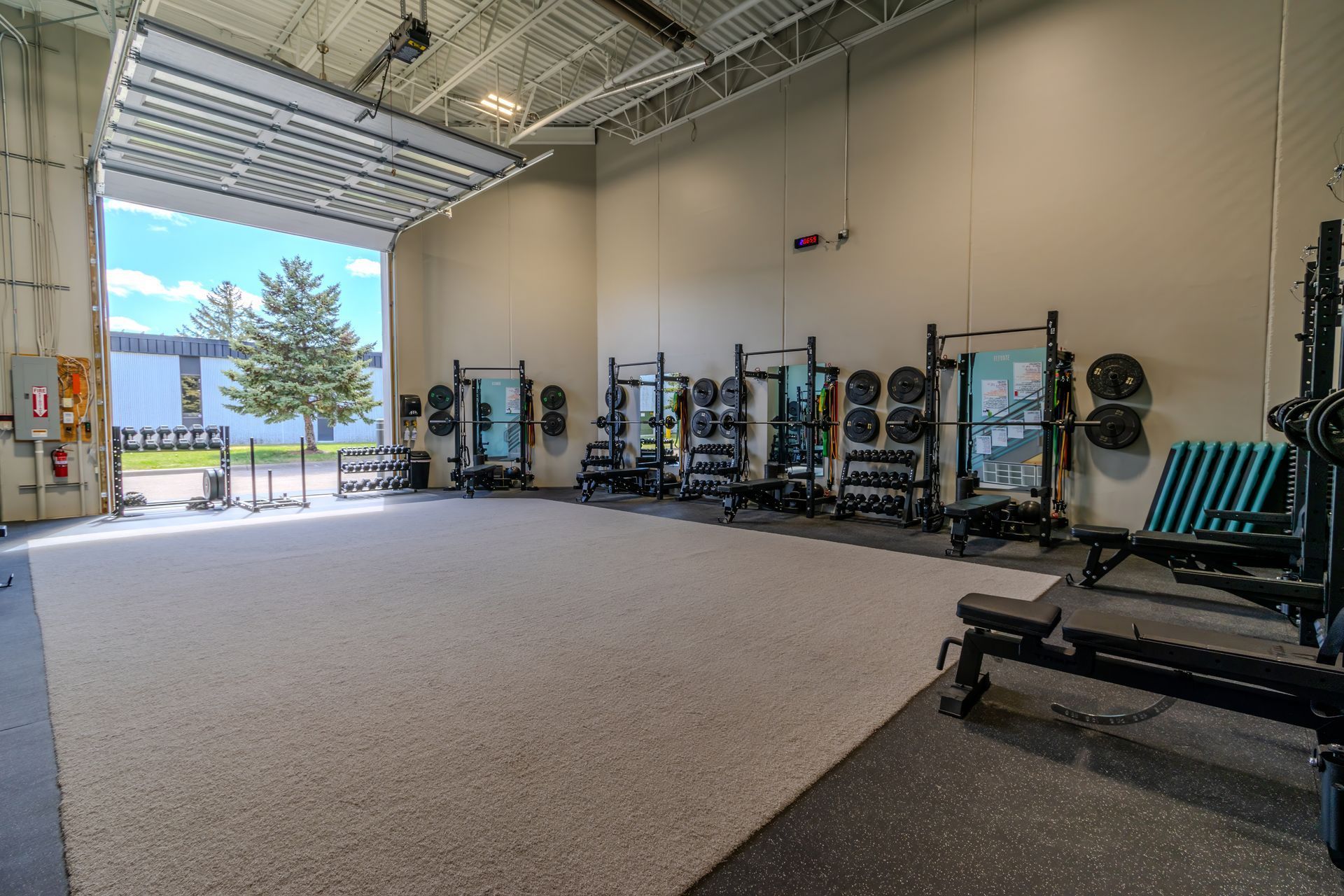 Empty gym with equipment-lined walls and an open garage door to a sunny outdoor view.