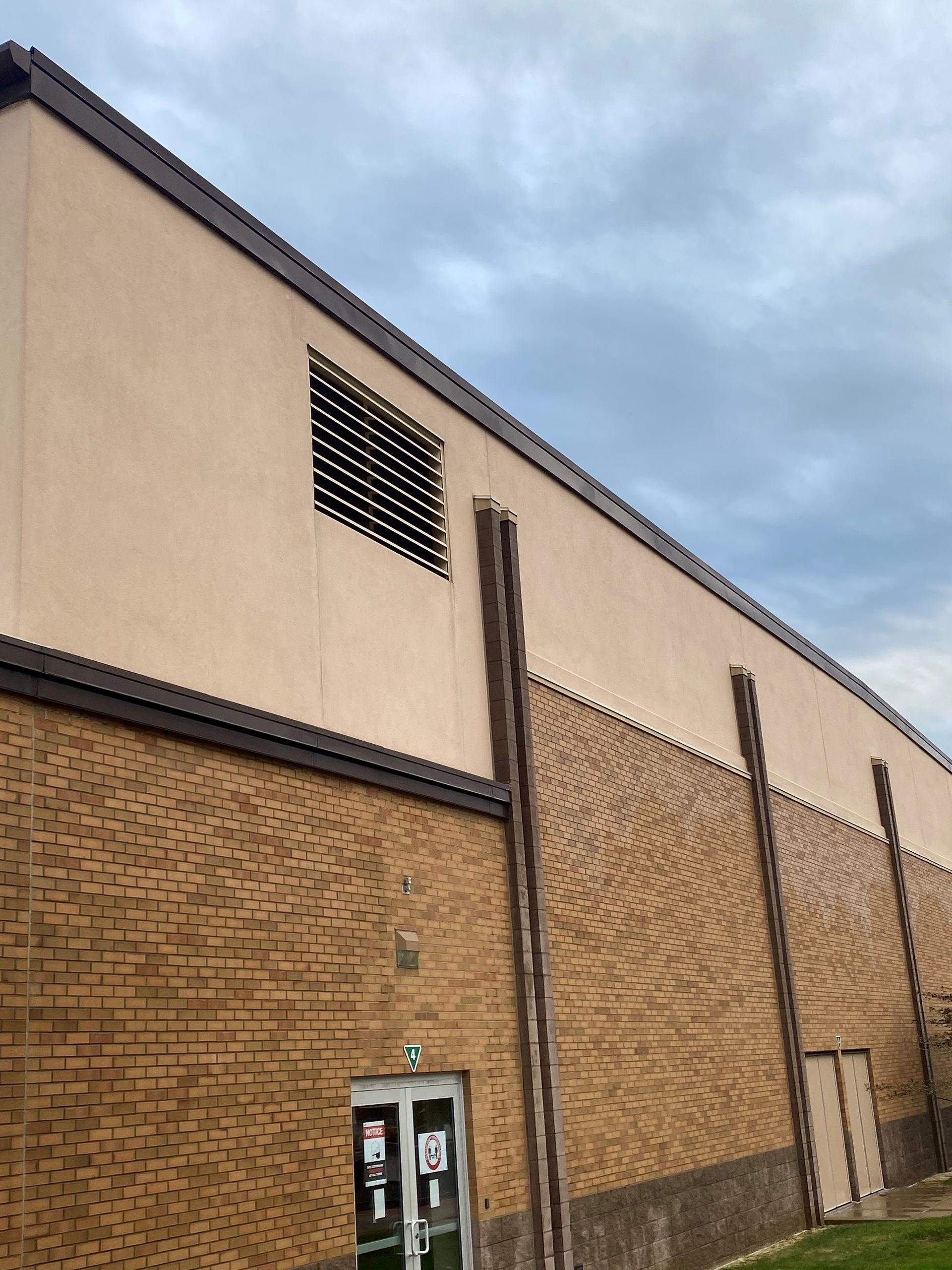 Tall tan brick building with a beige upper wall and a cloudy sky above the roofline