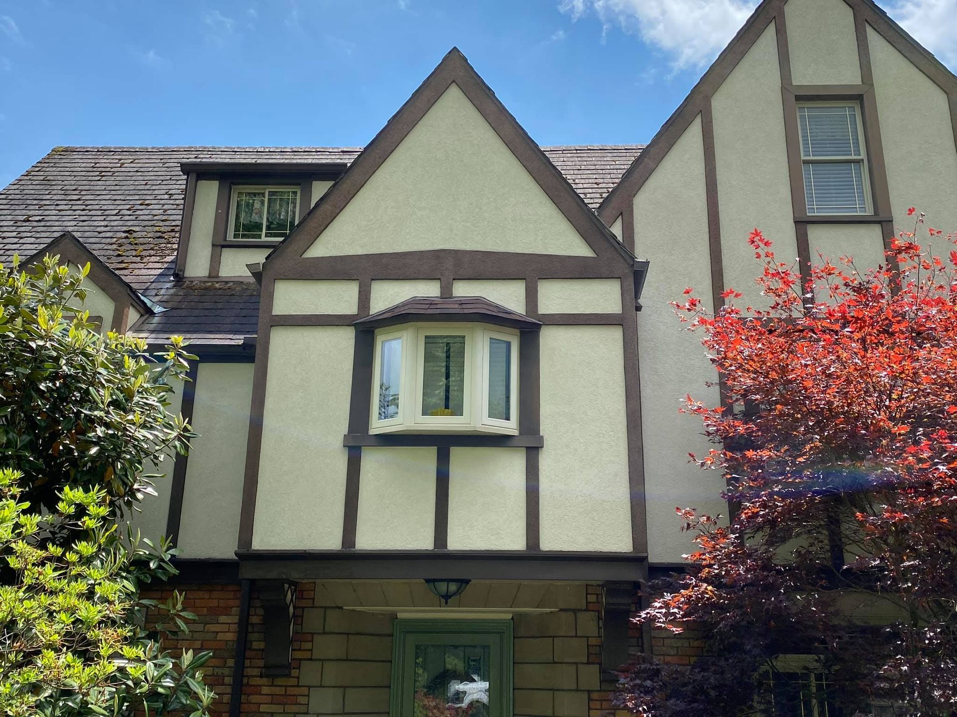 Tudor-style house facade with tan and dark trim, bay window, and trees on either side under a blue sky