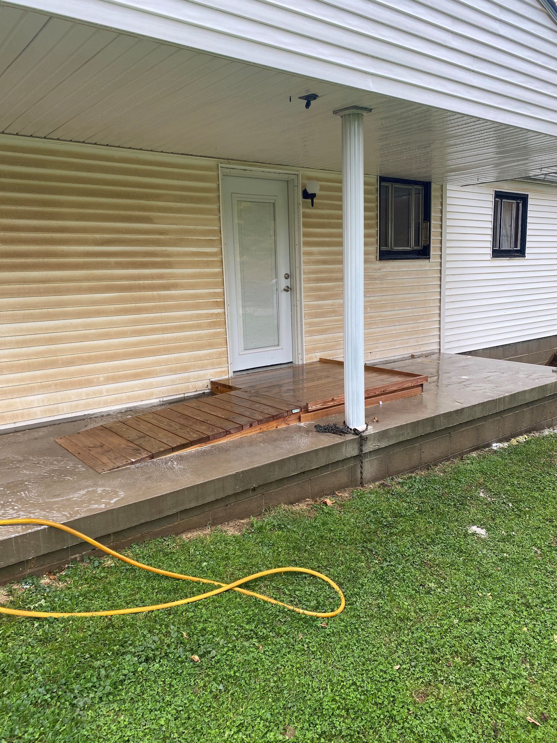 Front porch with white siding, pillars, closed door, windows, wet concrete, and a hose on the grass.