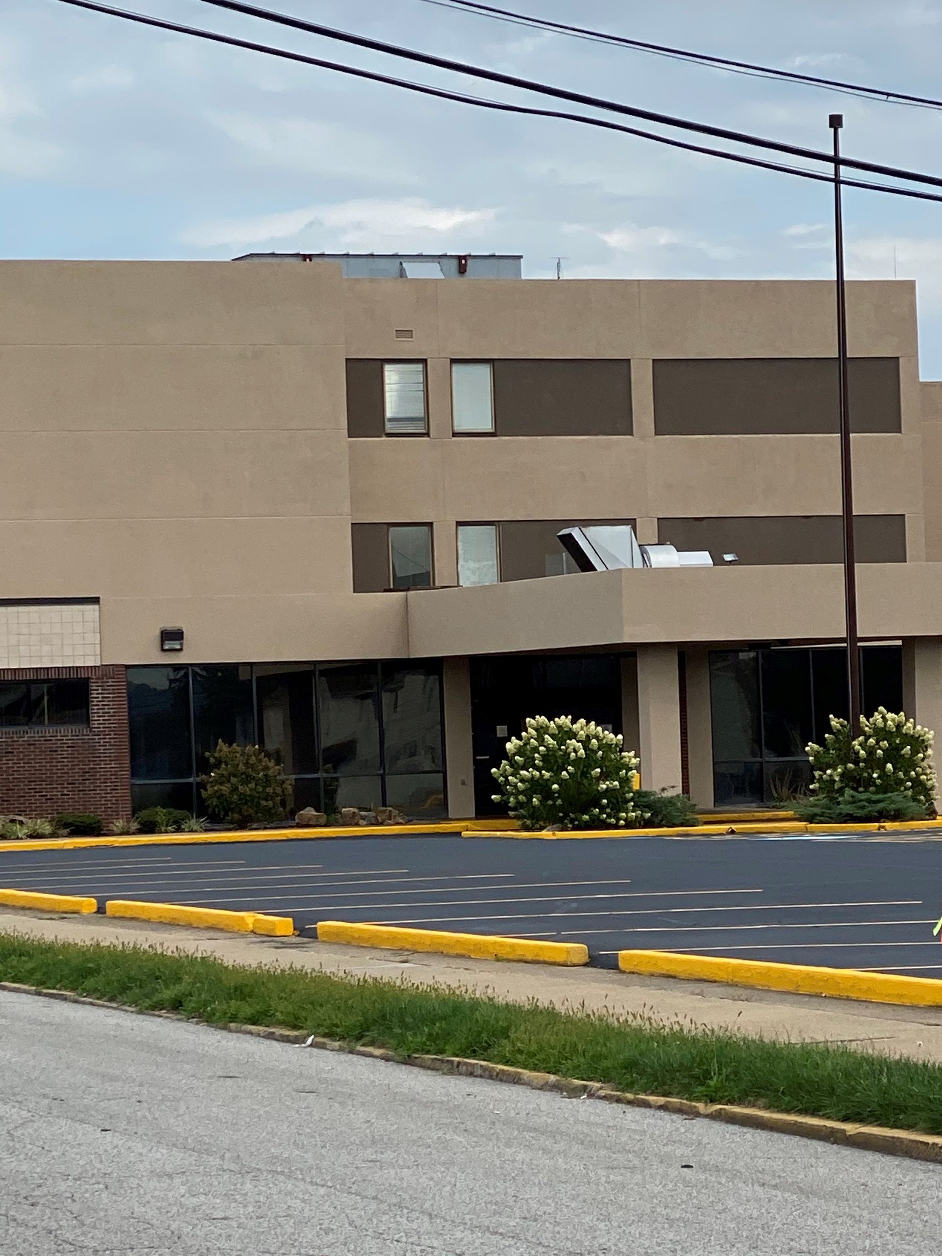 Tan beige commercial building with dark windows, shrubs, and a parking lot in front.