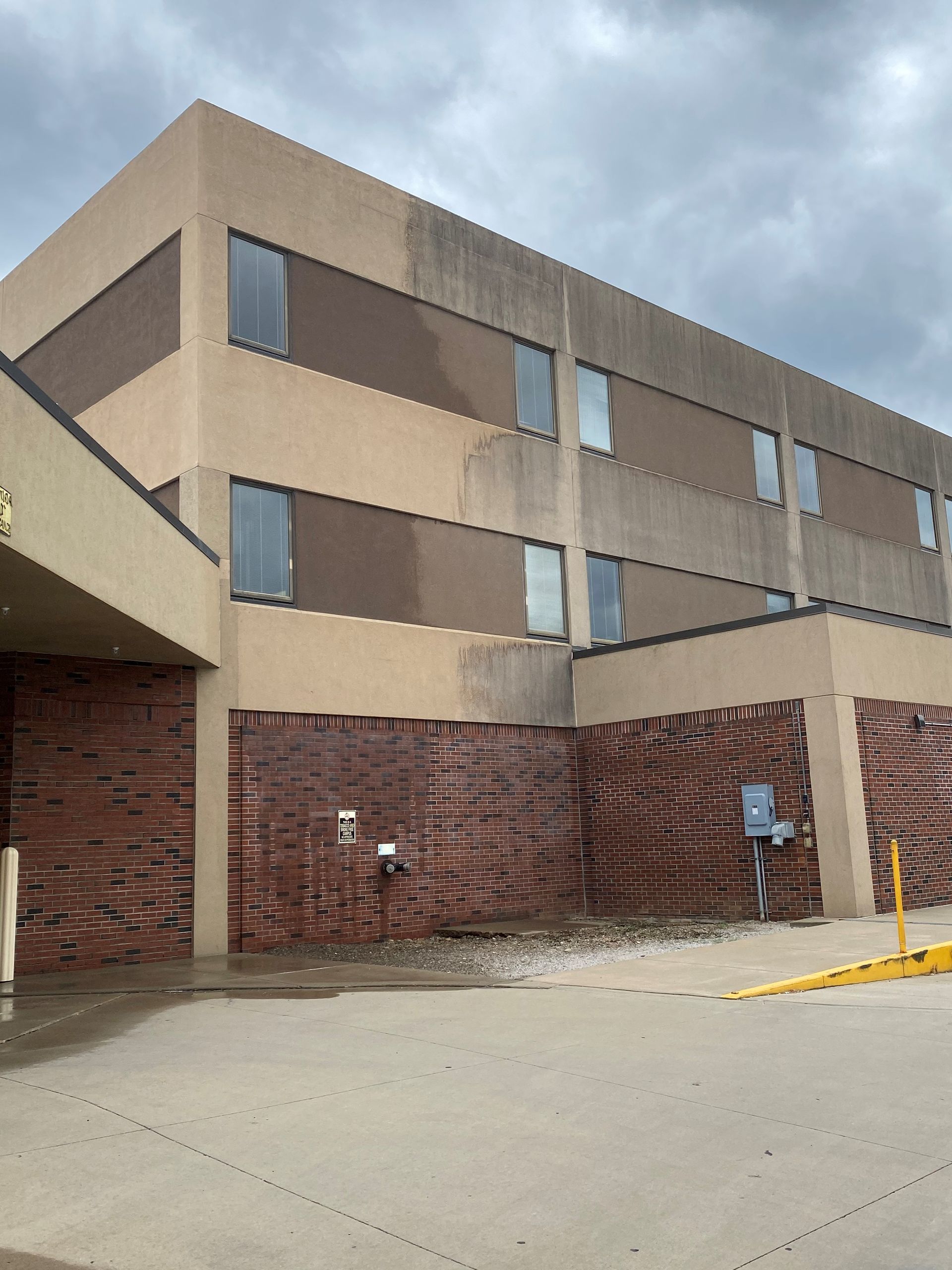Three-story tan brick building with a covered entrance and parking area under cloudy skies