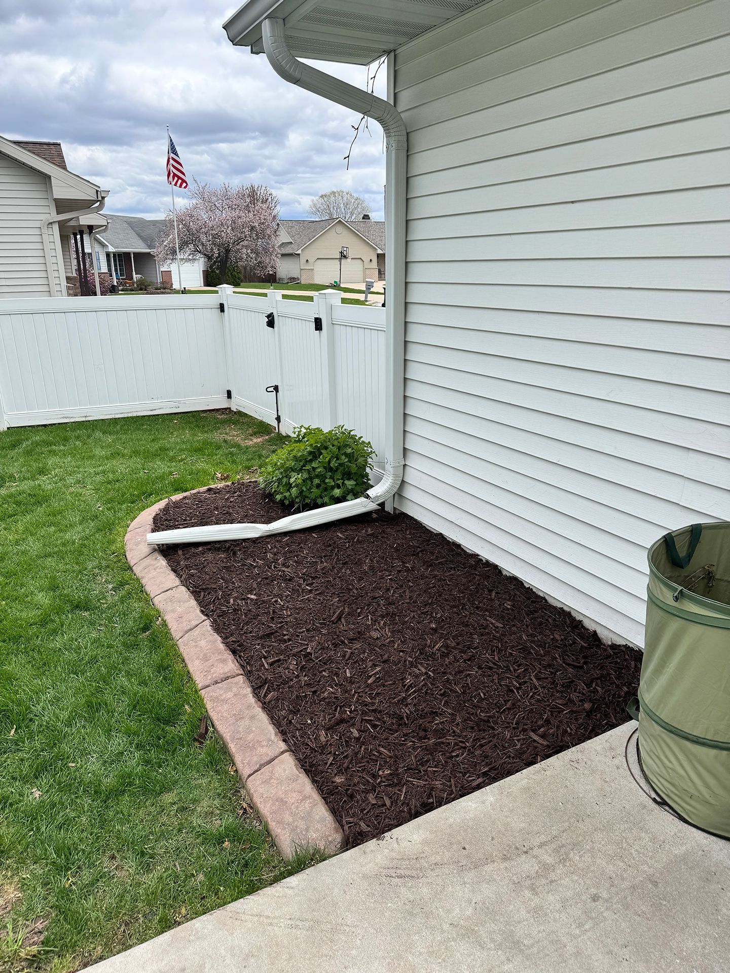 Backyard corner with white siding, mulched garden bed, grass, and vinyl fence near a concrete patio