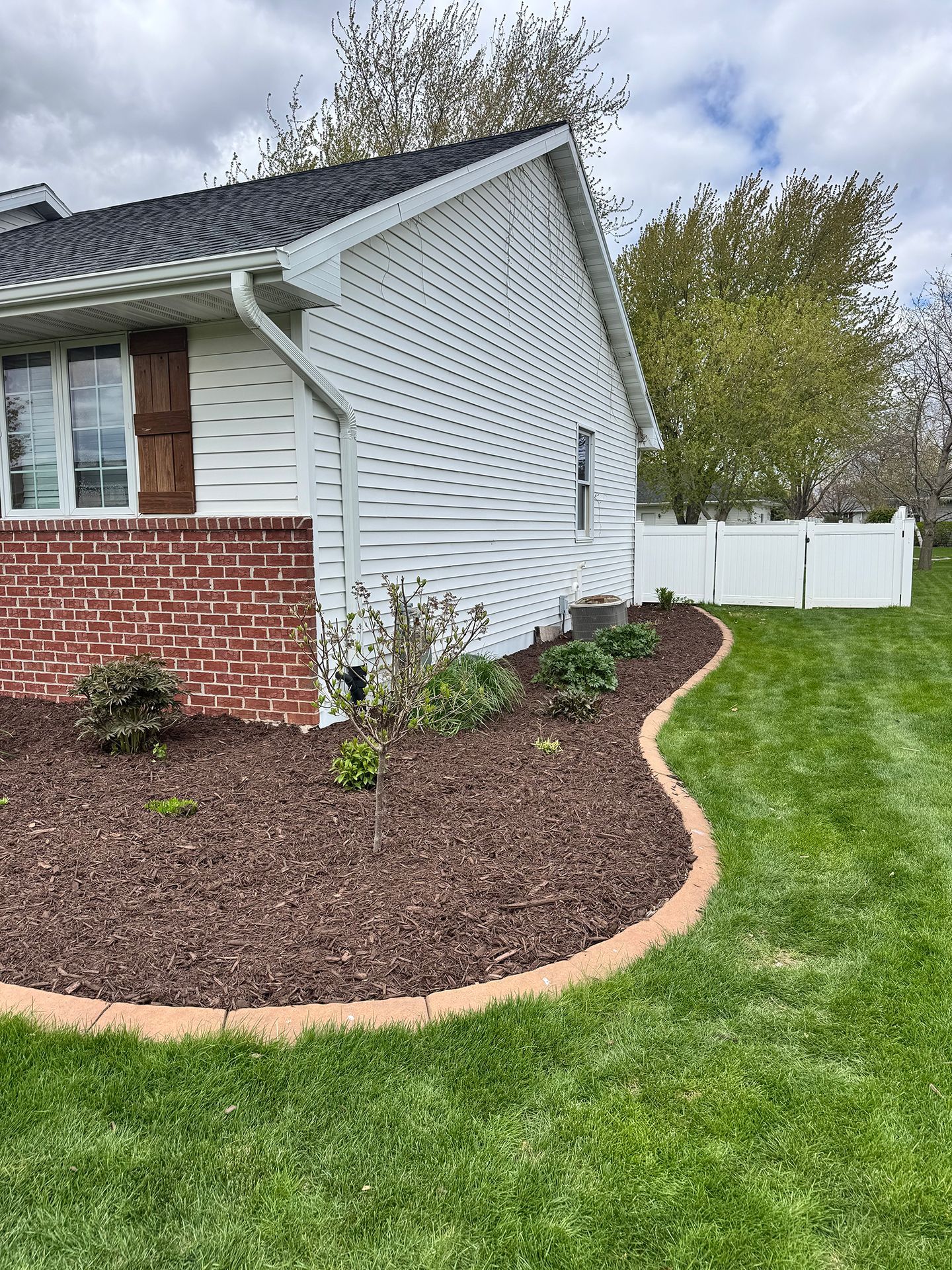 House corner with white siding, brick foundation, curved mulched bed, and green lawn by a white fence