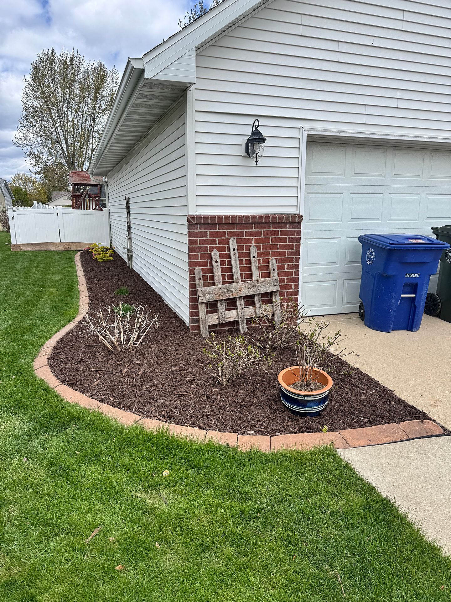 Side yard with brick-and-vinyl house corner, garage, mulch bed, small trellis, and blue trash bin