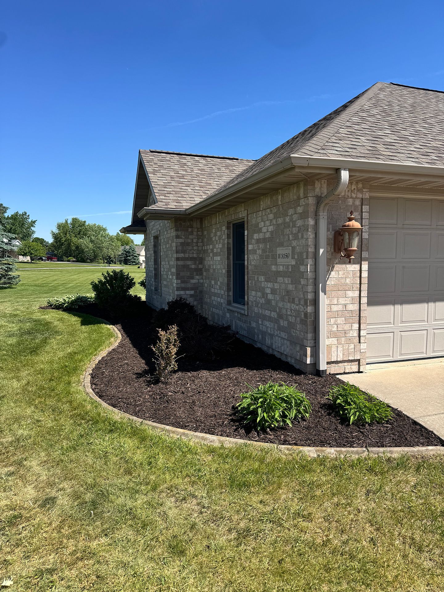 Brick house corner with garage, lantern, and mulched shrubs beside a green lawn under a clear blue sky