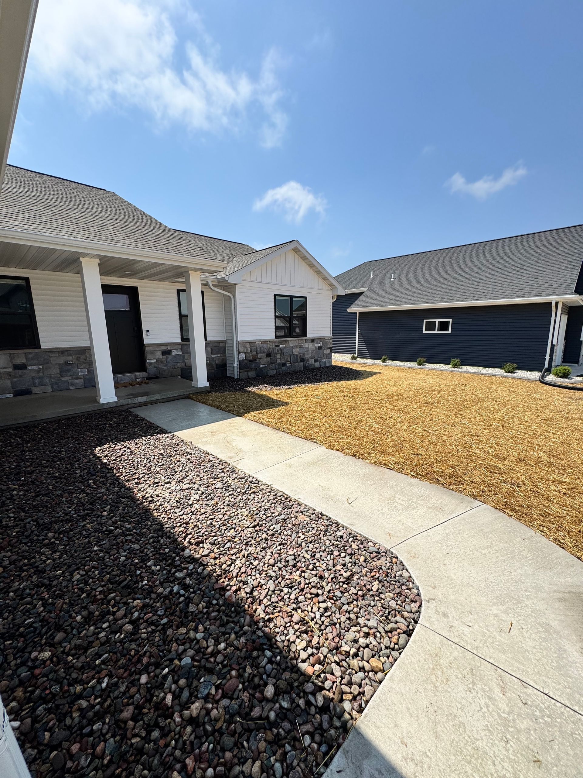 Curved concrete walkway between dark mulch and tan gravel leading to a white house under a blue sky