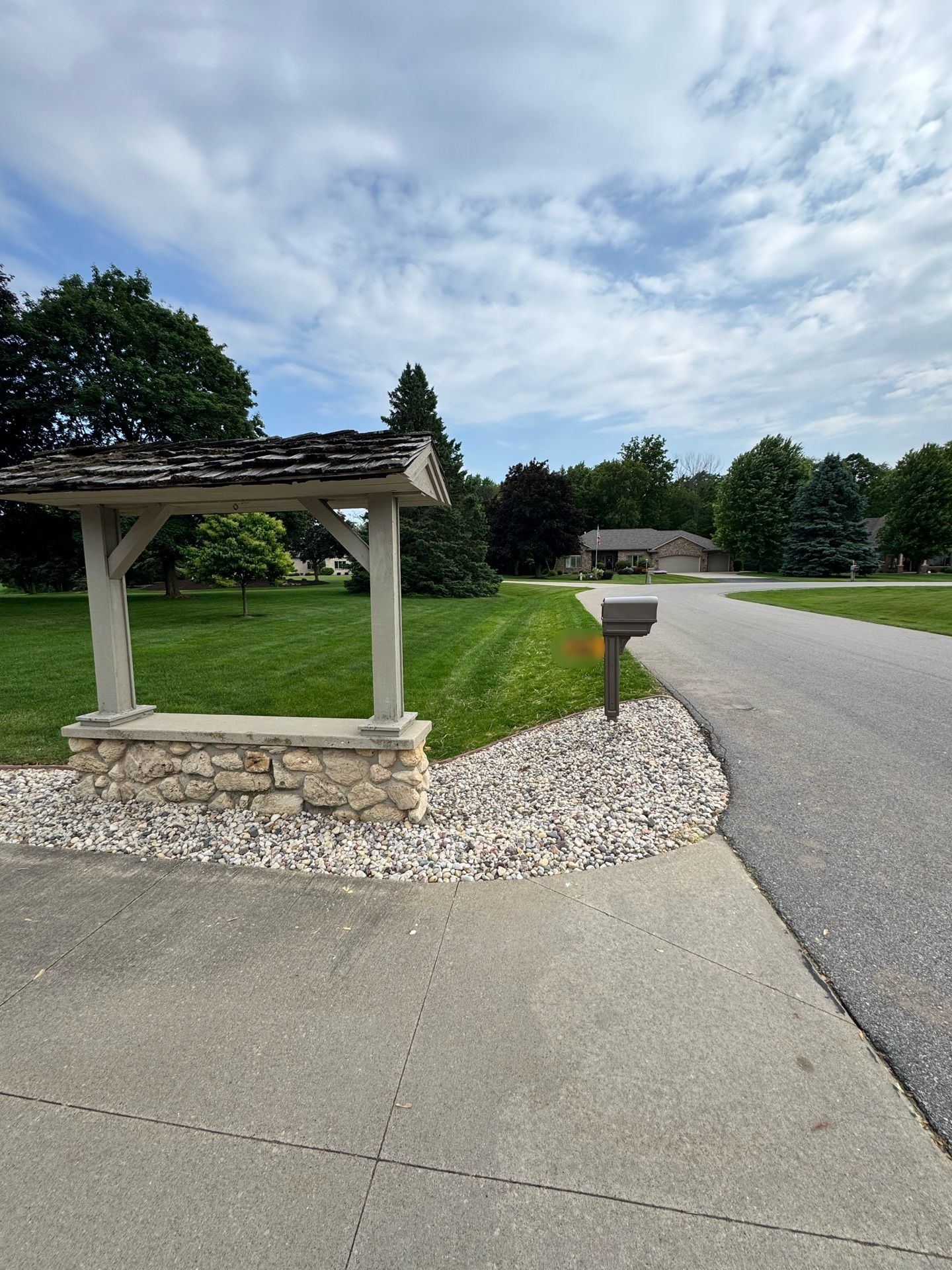 Stone mailbox and pergola beside a driveway in a grassy suburban neighborhood under a cloudy sky