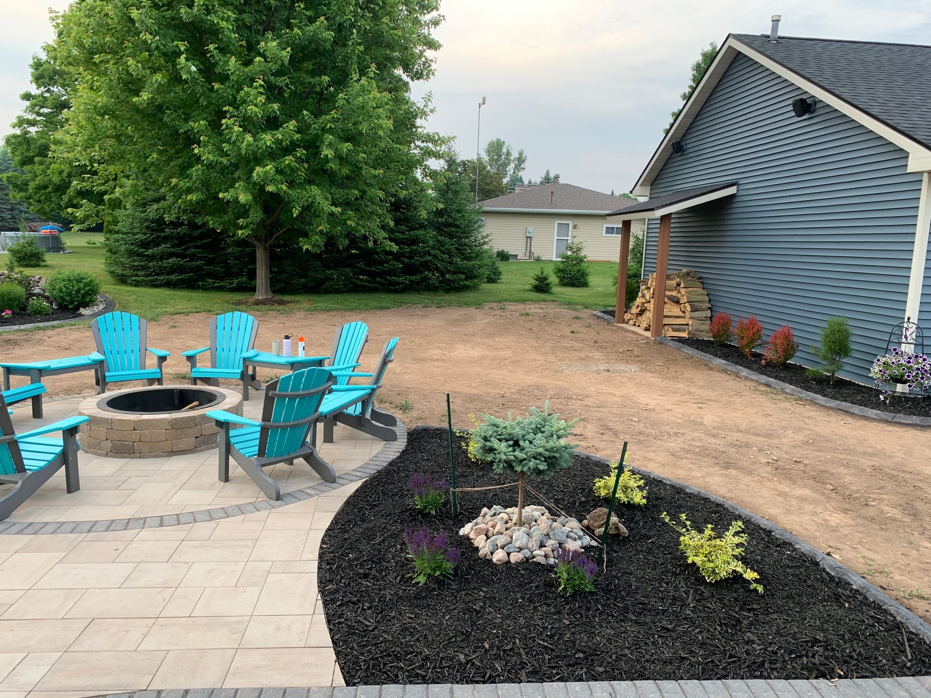 Backyard patio with turquoise chairs around a fire pit, landscaped mulch beds, and a blue house exterior