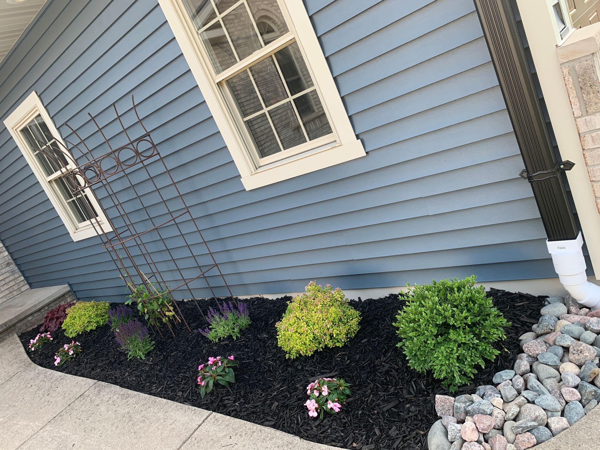 Blue house siding with small landscaped flower bed, shrubs, mulch, and decorative rocks along the foundation