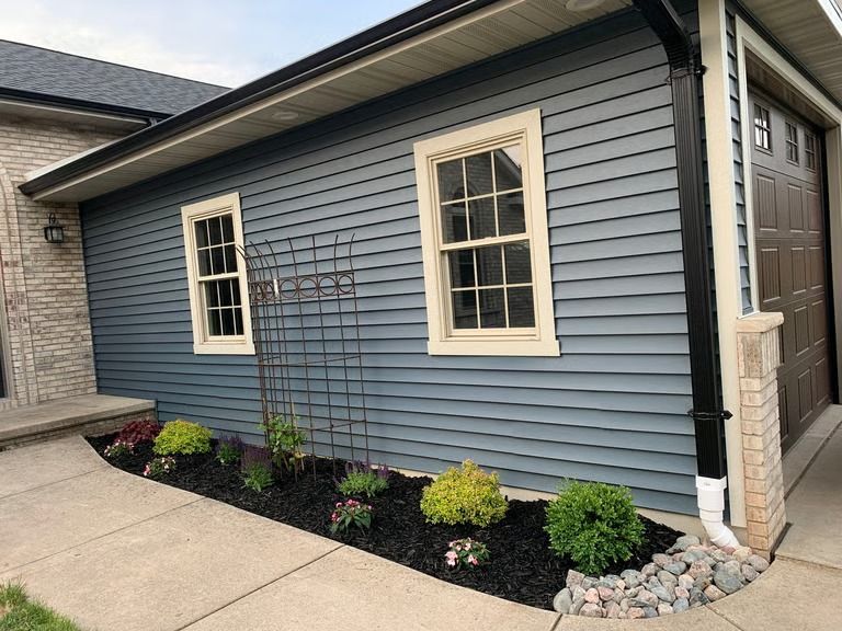 Blue house exterior with white-trimmed windows, concrete walkway, and small landscaped flower bed