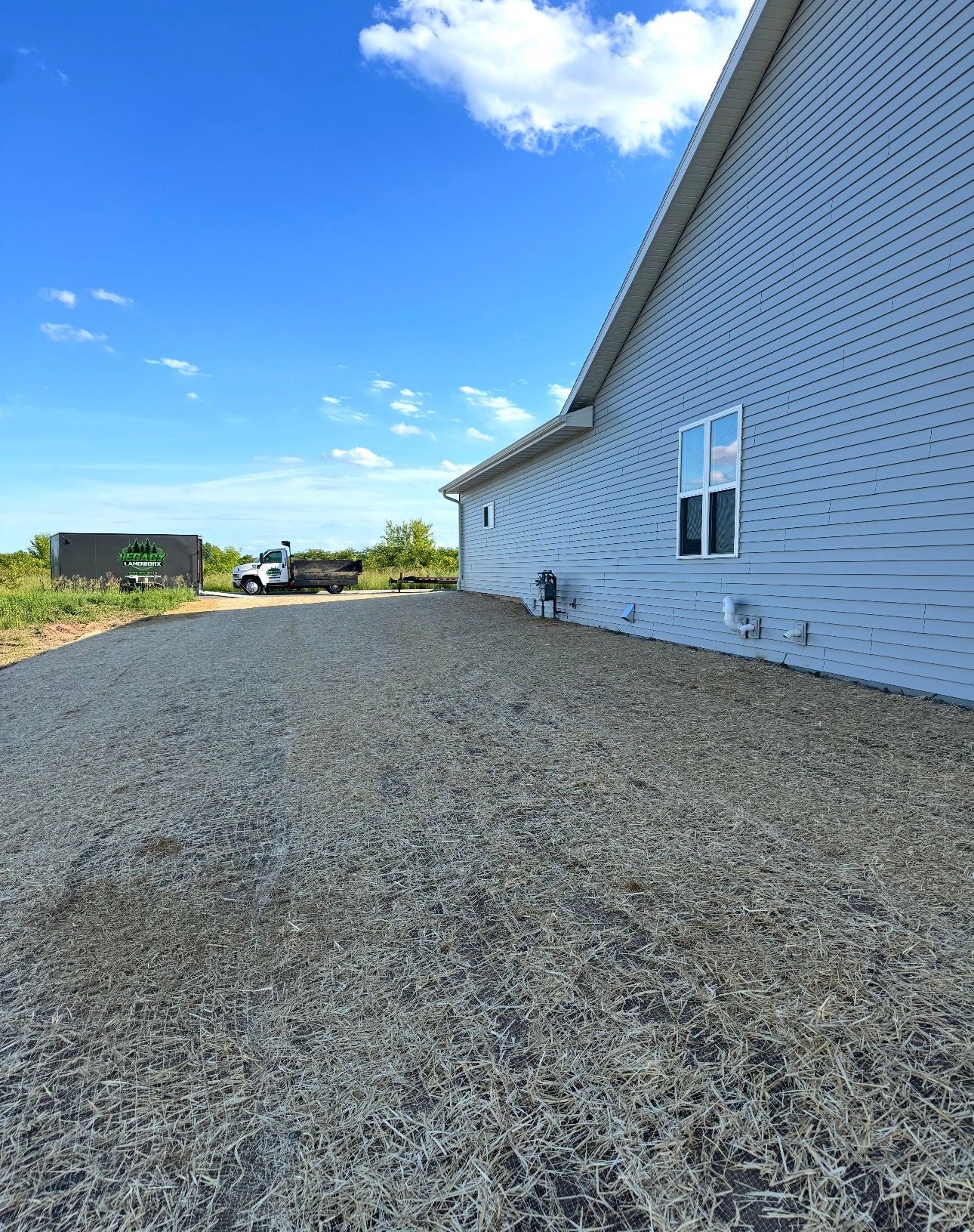 Gravel driveway beside a white house under a bright blue sky with scattered clouds.