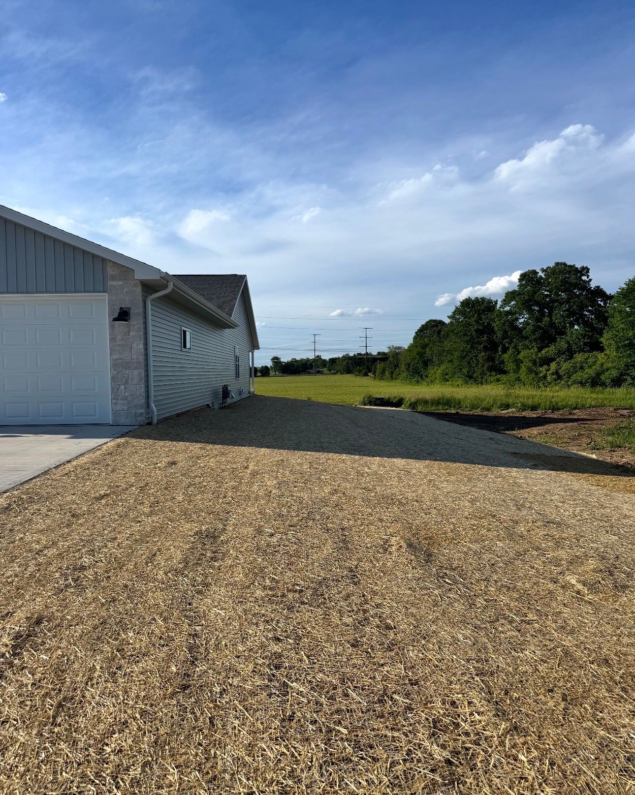 Gravel driveway beside a white house under a blue sky, with trees and open land in the distance