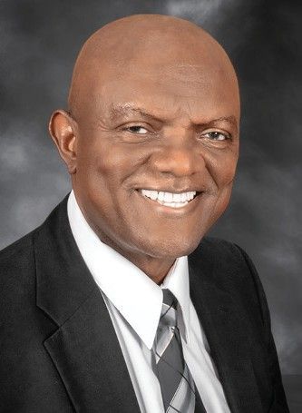 Portrait of a smiling bald man in a black suit, white shirt, and striped tie on a gray background