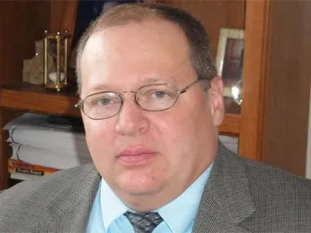 Man in glasses and gray suit seated indoors in front of wooden shelves