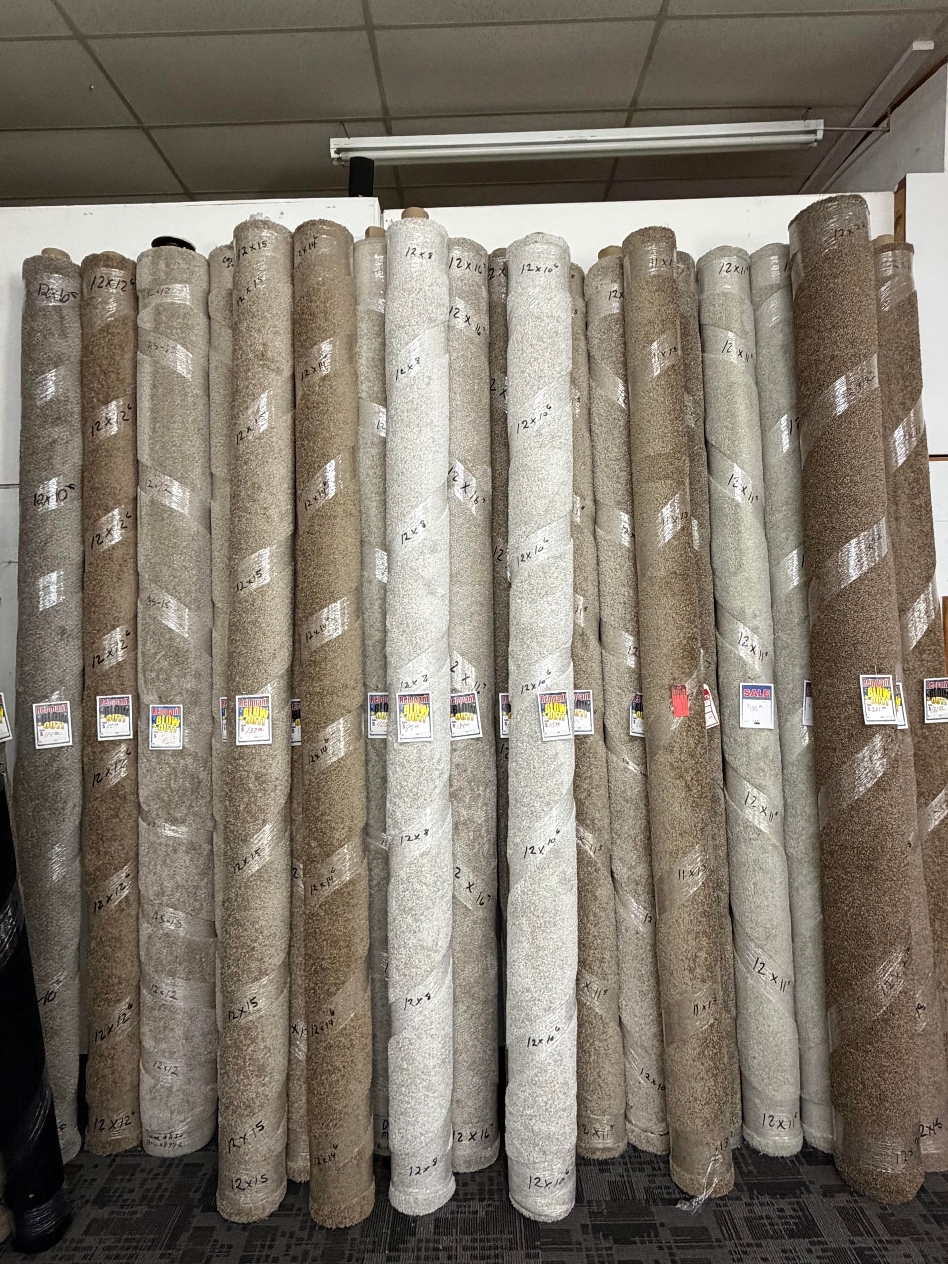 Rolled carpet samples stacked upright in a store aisle, in shades of gray, beige, and brown.