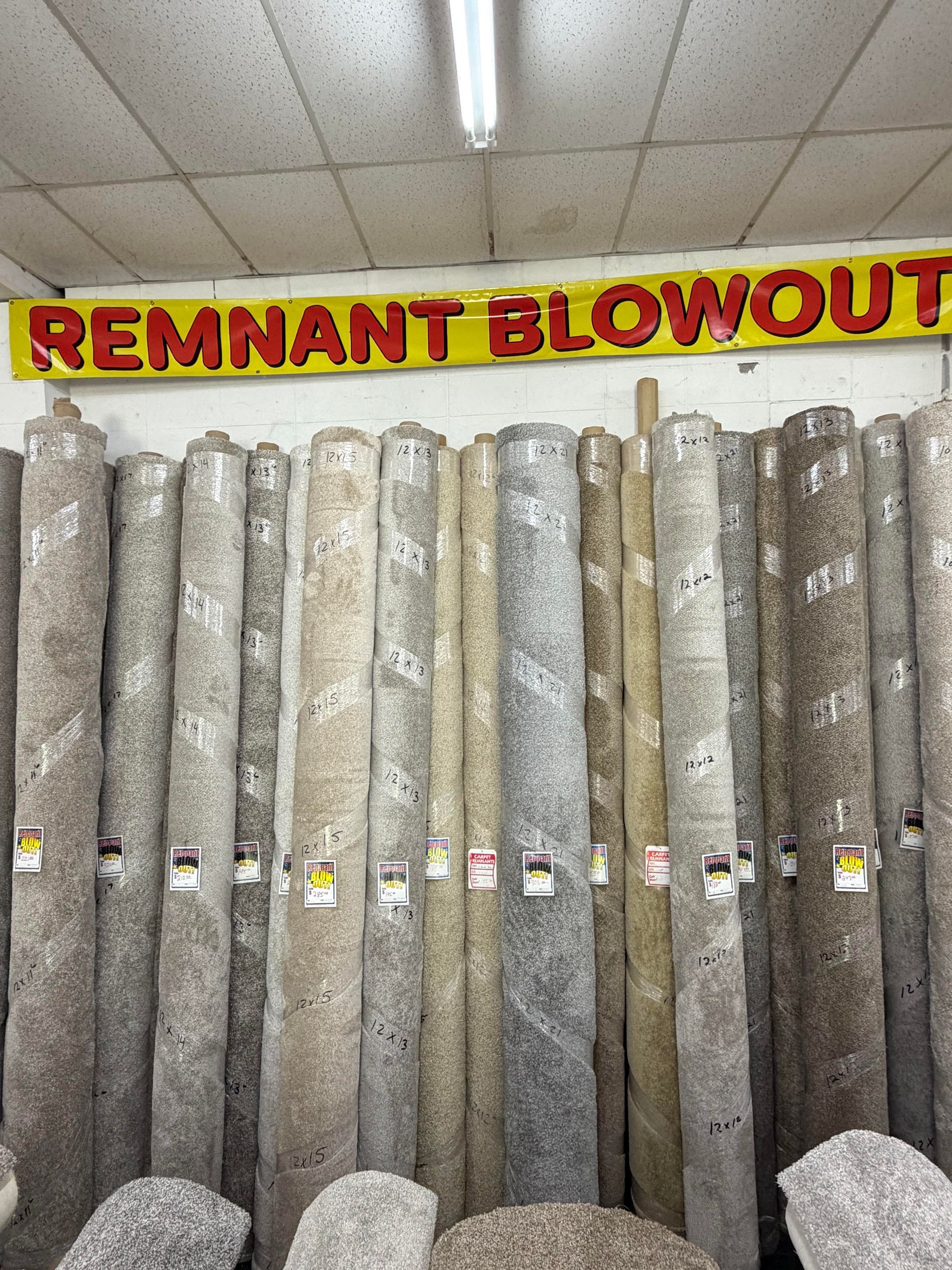 Carpet rolls stacked upright beneath a red-and-yellow “REMNANT BLOWOUT” store sign.