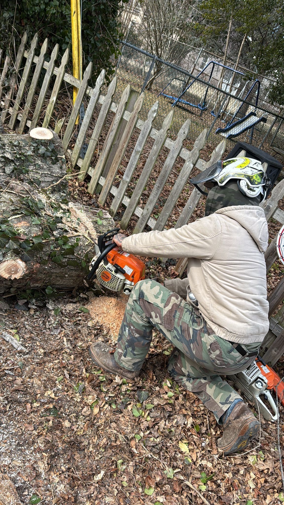 Person in safety gear using chainsaw to cut a log near a fence.